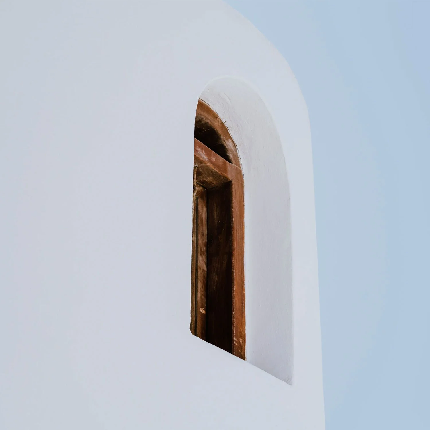 Close-up of a small, arched window with a wooden frame, set into a white wall.