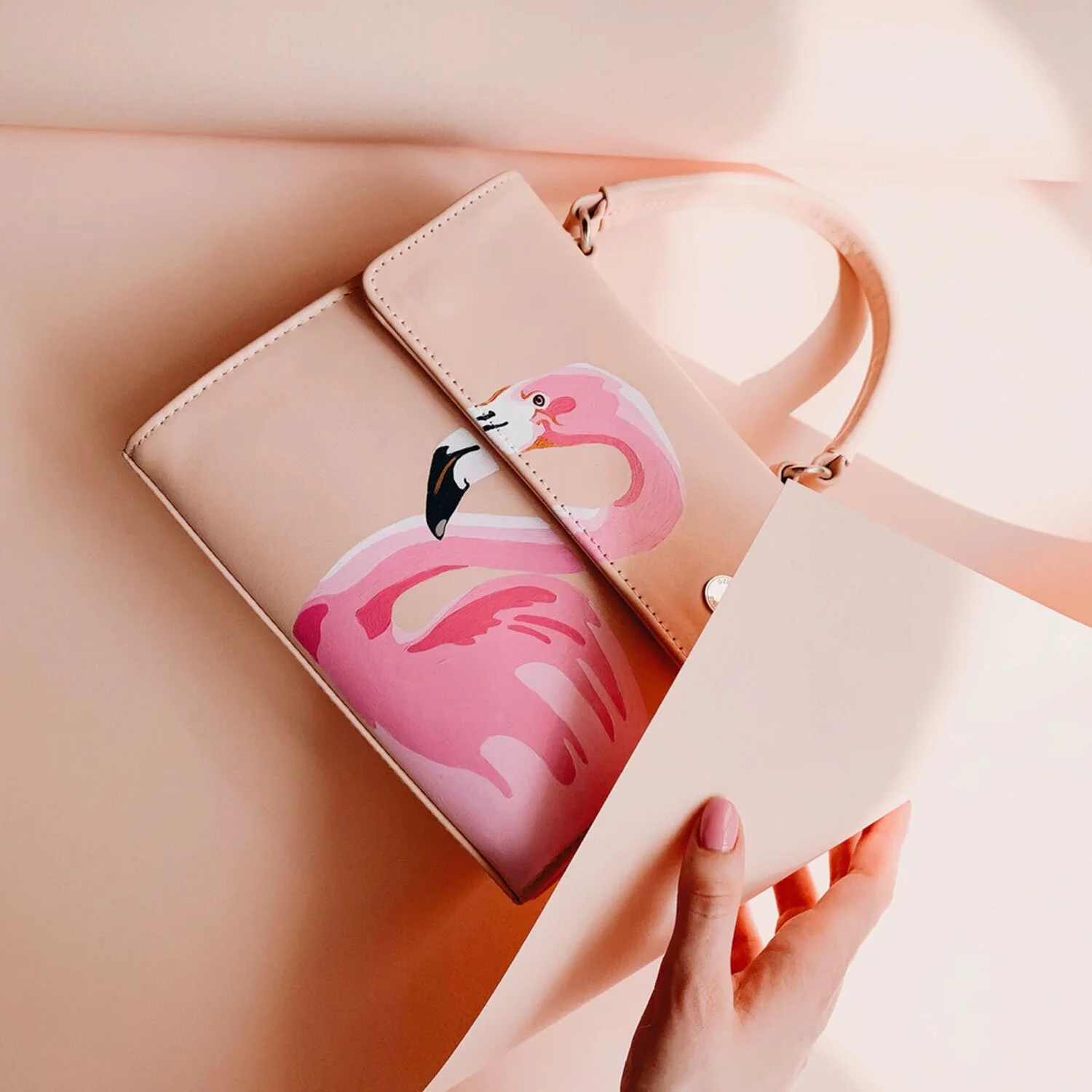 A pink handbag with a flamingo illustration inside a slightly opened pink box, with soft pink background.