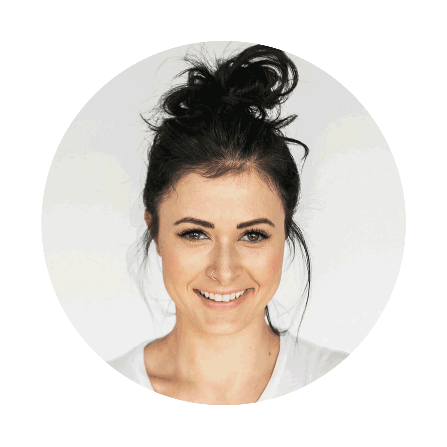 A young woman with dark hair styled in a messy bun, smiling, with a nose piercing, wearing a white top, against a white background.