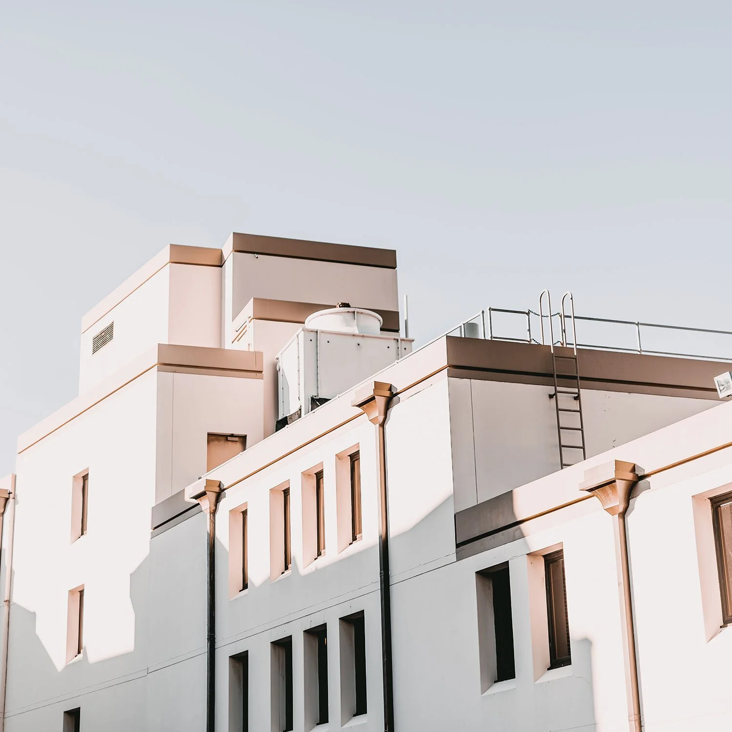 The top portion of a modern white apartment building with pink accents, city windows, a ladder, and rooftop equipment under a clear sky.
