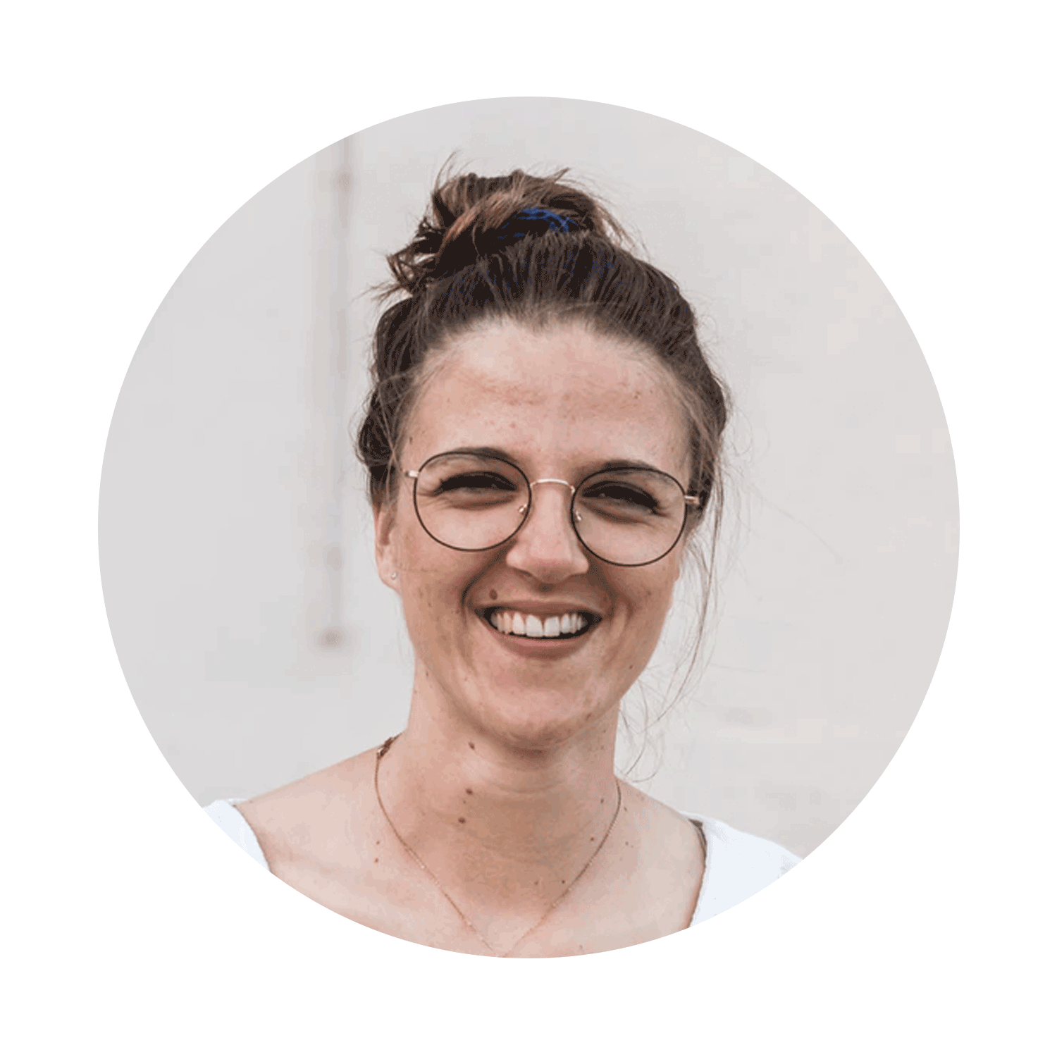 A smiling woman with glasses and a bun hairstyle, wearing a white top, standing against a plain light background.