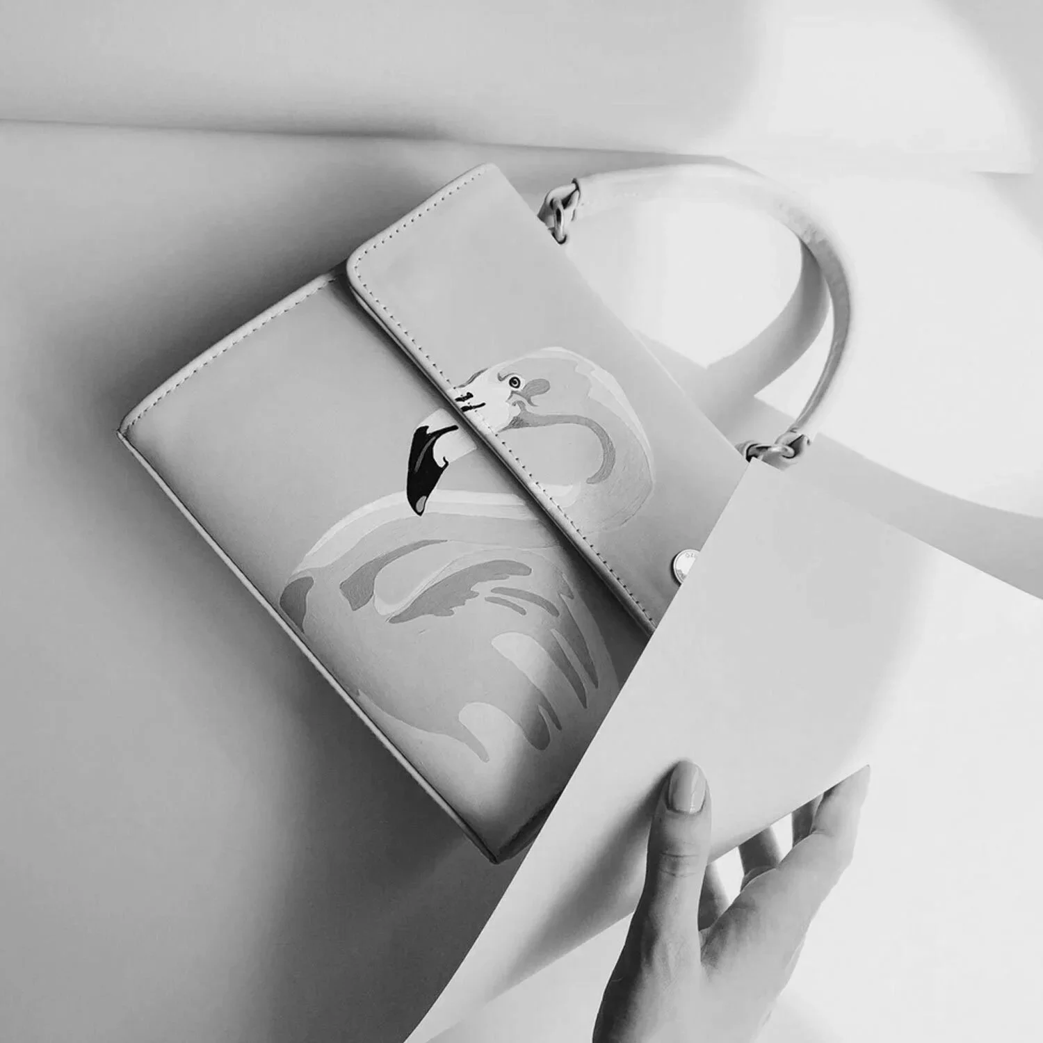 Black and white photograph of a hand holding a paper bag with a flamingo illustration, outside a shopping bag with a white purse resting on top.