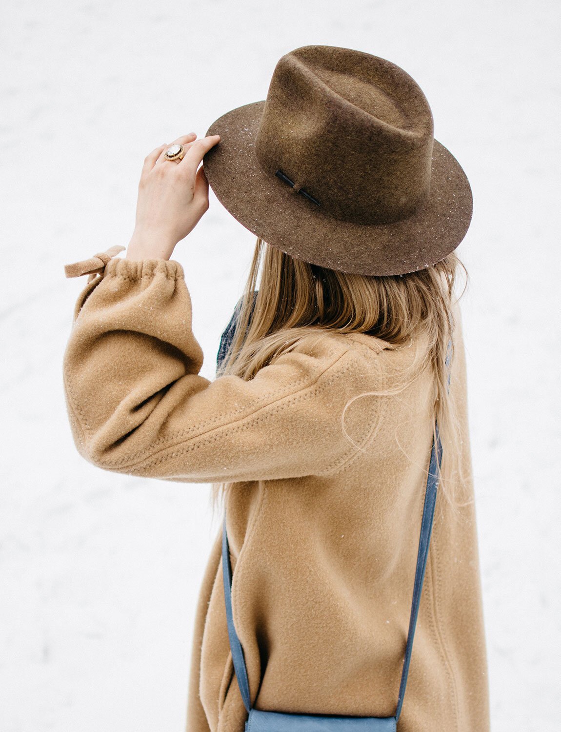 Person wearing a brown hat and camel-colored coat, standing outdoors in a snowy setting, with their hand adjusting the hat.