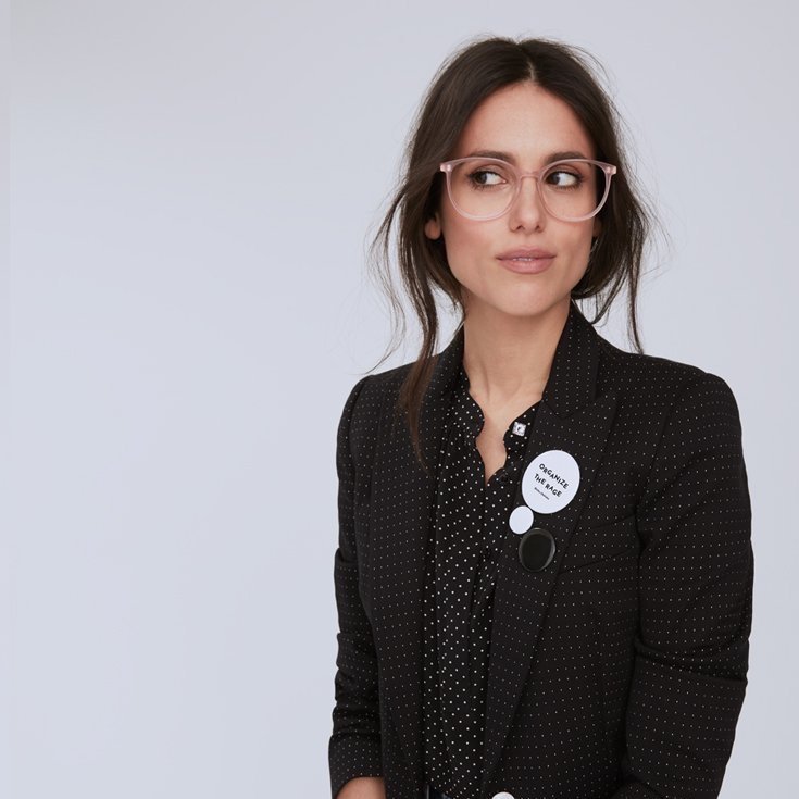  Liz Plank, a woman wearing glasses with brown hair pulled back into a bun, looking off to the side, wearing a black dotted blazer over a black dotted blouse 