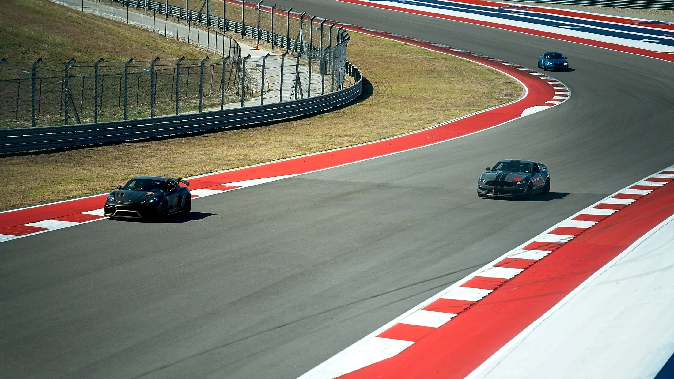 Three sports cars on a race track, two black and one blue, navigating a curve with red and white painted curbing.