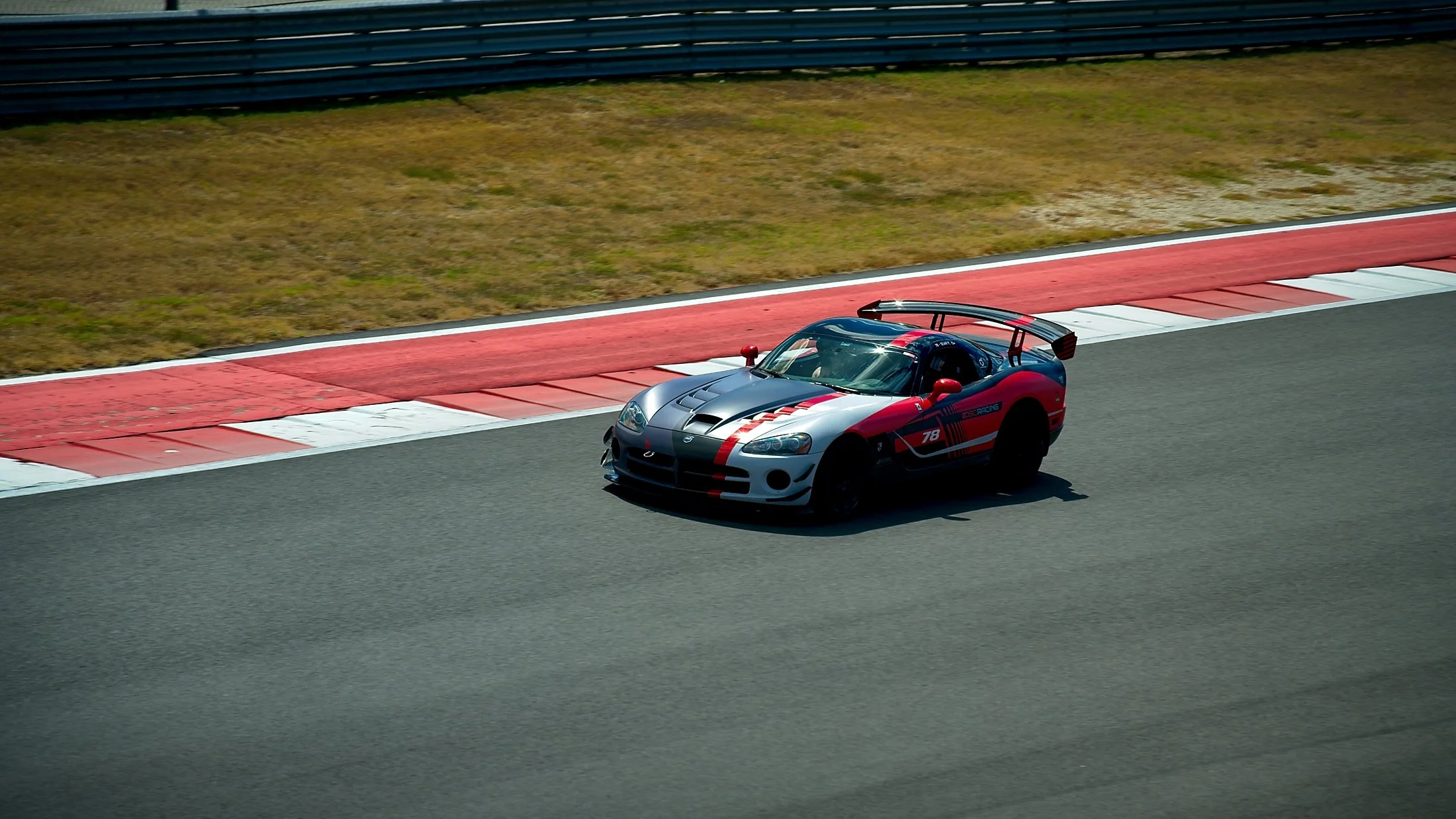 A silver and red race car driving on a asphalt track with red and white curbing, in an open race circuit with grass and barriers in the background.