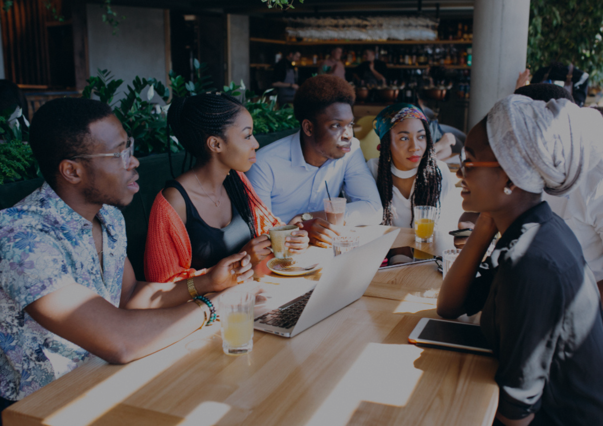 Group of five young professionals collaborating around a laptop at a café table with drinks, engaged in an informal discussion.