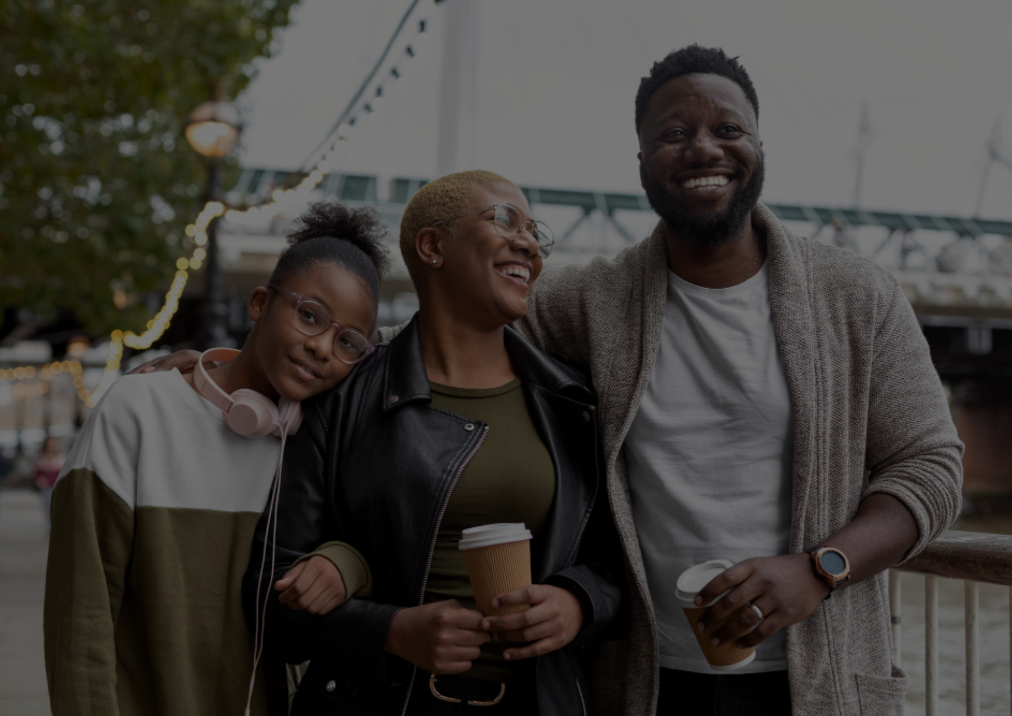 Family of three smiling outdoors on a city walkway holding coffee cups together.