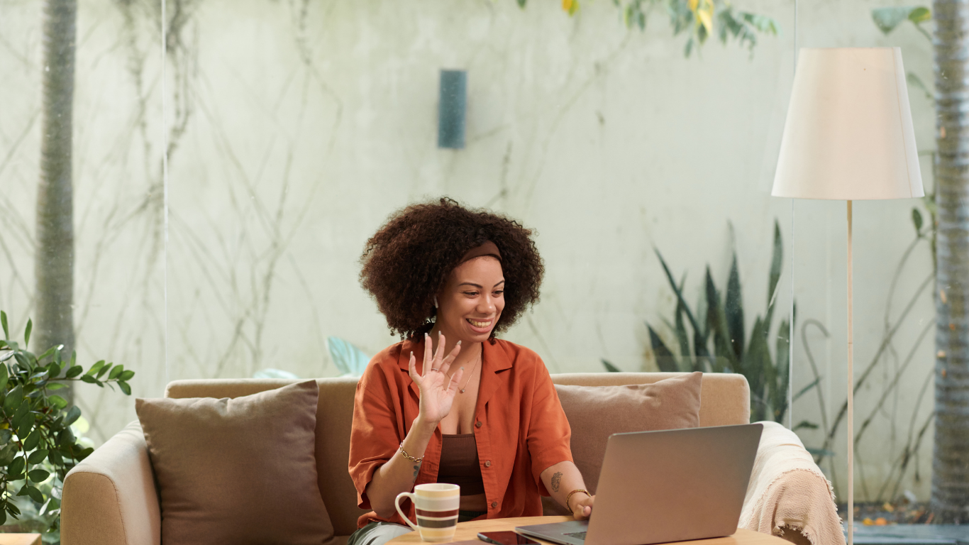 Woman smiling and waving during a virtual meeting on her laptop while seated on a couch with a coffee mug.