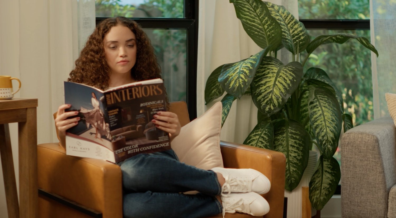A young woman with curly hair sitting on a tan leather armchair reading a magazine titled 'Interiors'. She's wearing white sneakers, ripped jeans, and a white top. There is a large leafy plant next to her and a window in the background.