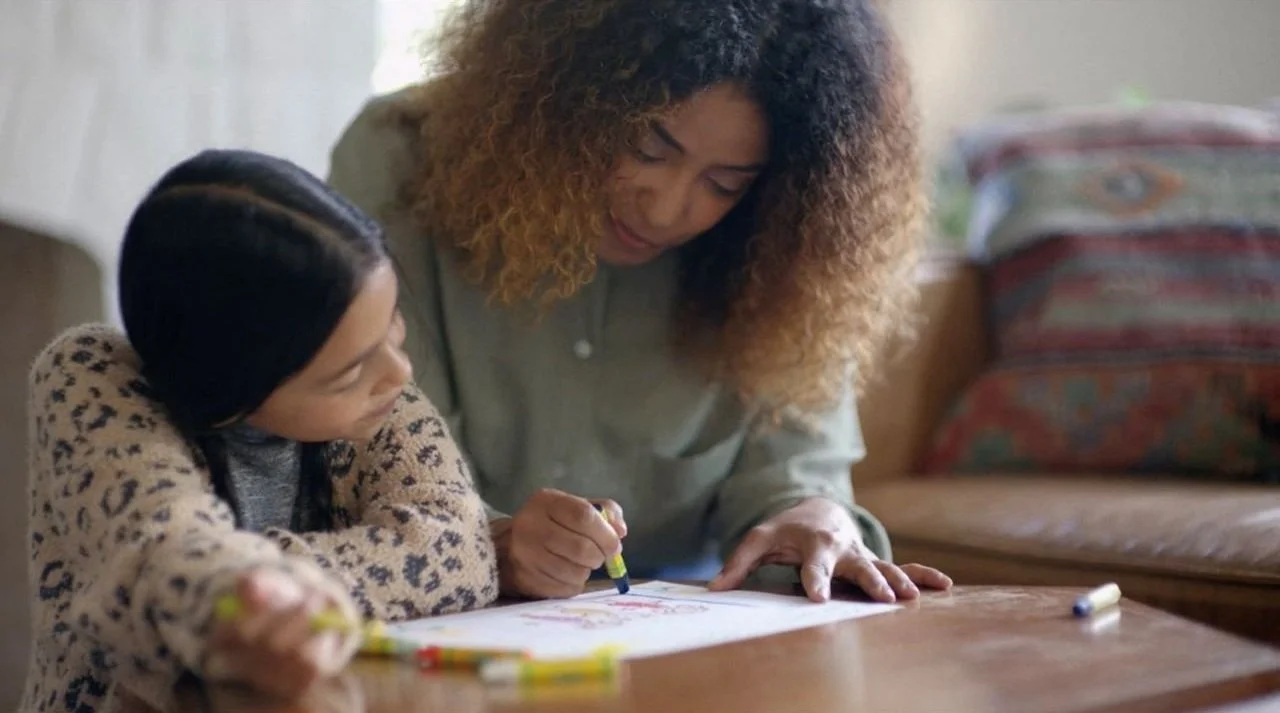 A woman and a young girl drawing together at a wooden table in a cozy room.