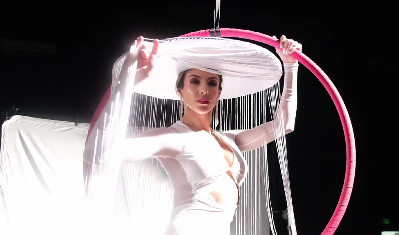 A woman with dark hair and makeup, dressed in white, holds a large pink and white hoop with a matching white and pink hat, with hanging white strings, against a dark background.
