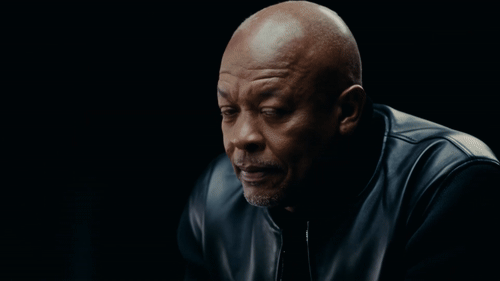 Close-up of a middle-aged man with a shaved head wearing a black leather jacket, looking down contemplatively against a dark background.