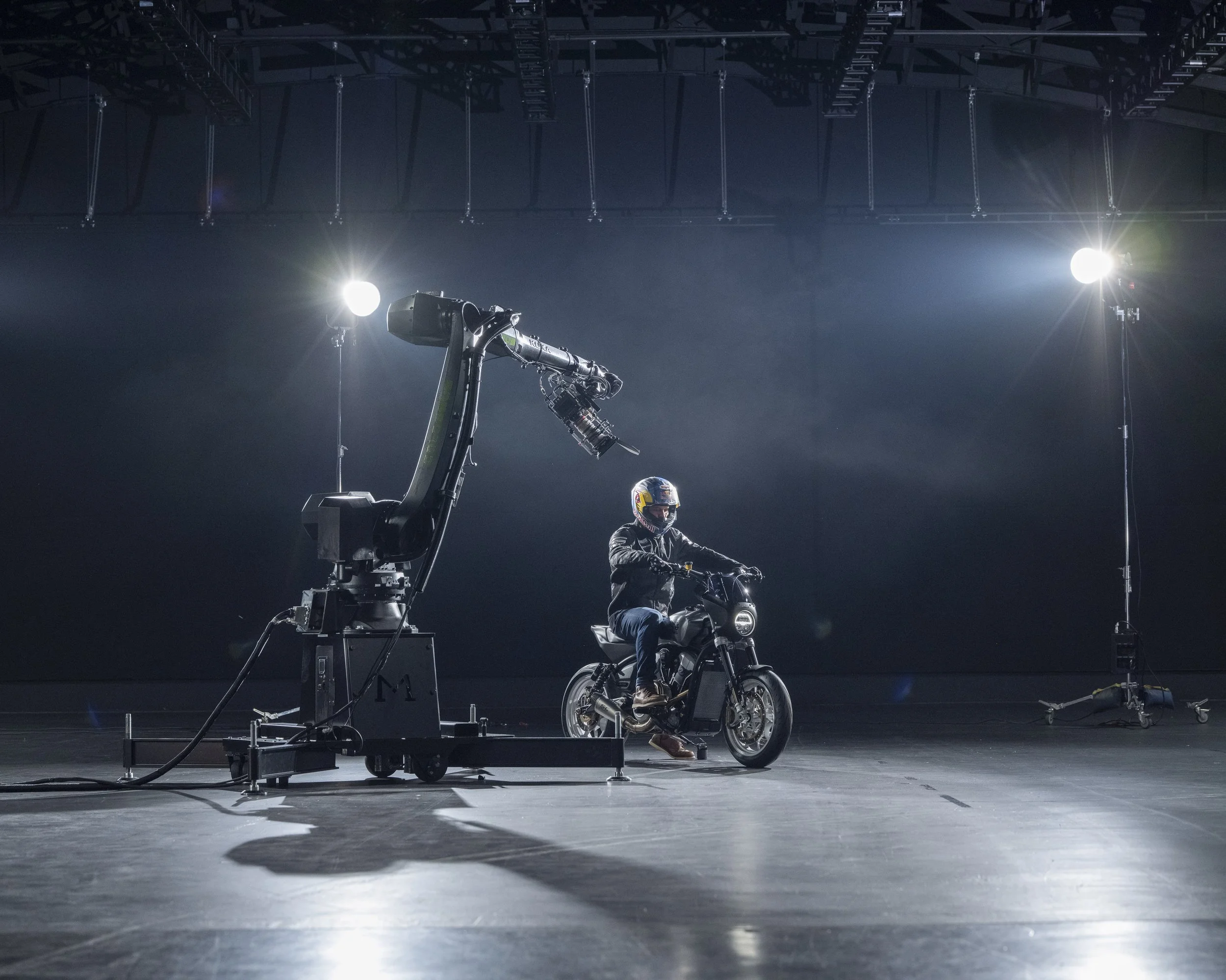 A person wearing a helmet and black jacket riding an electric motorcycle in a studio with professional lighting and a robotic arm nearby.