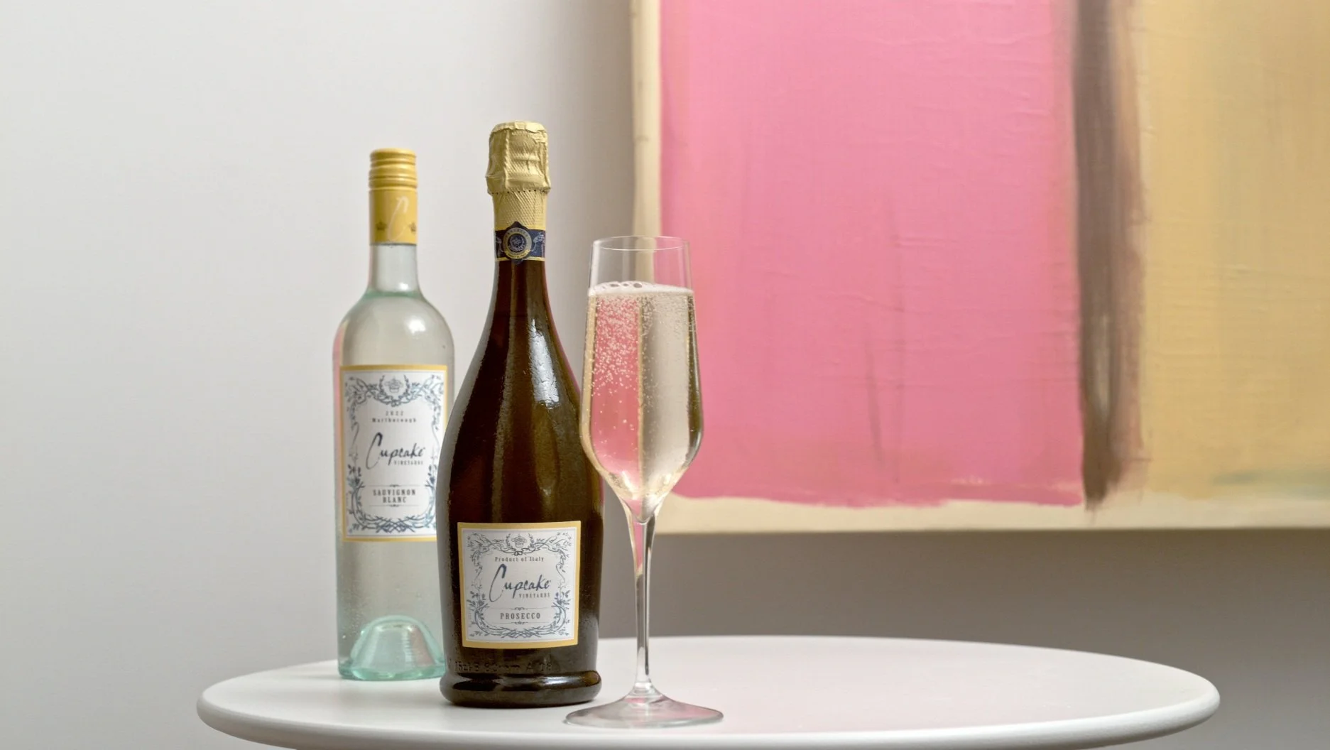Two bottles of wine, one white and one rosé, and a filled champagne flute on a round white table in front of an abstract pink and beige painting.