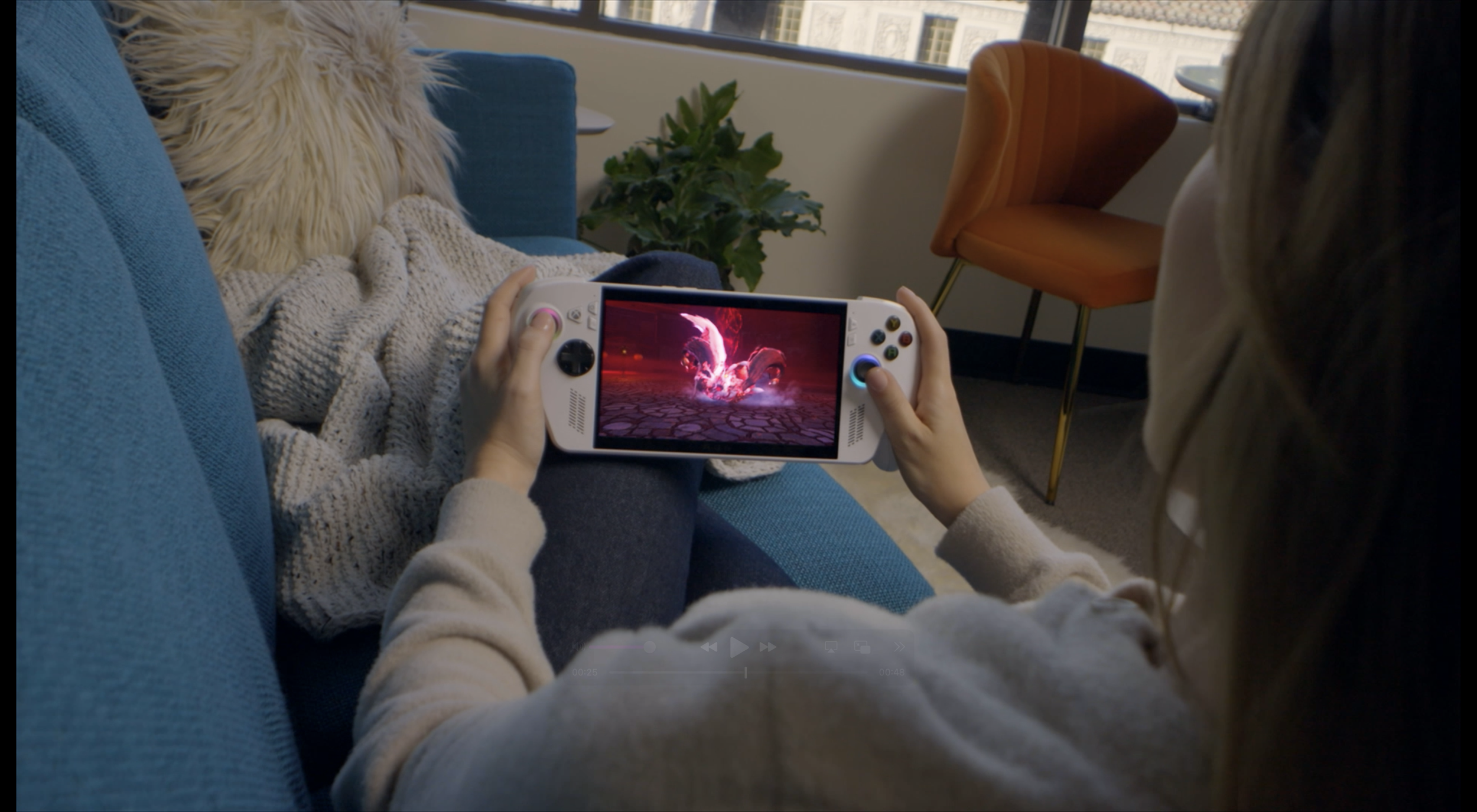 Person playing a video game on a handheld gaming device while sitting on a blue couch in a cozy living room.