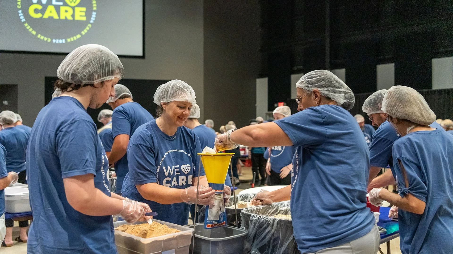 People wearing blue T-shirts and hairnets preparing food at a large community event, with a screen that says 'WE CARE' in the background.