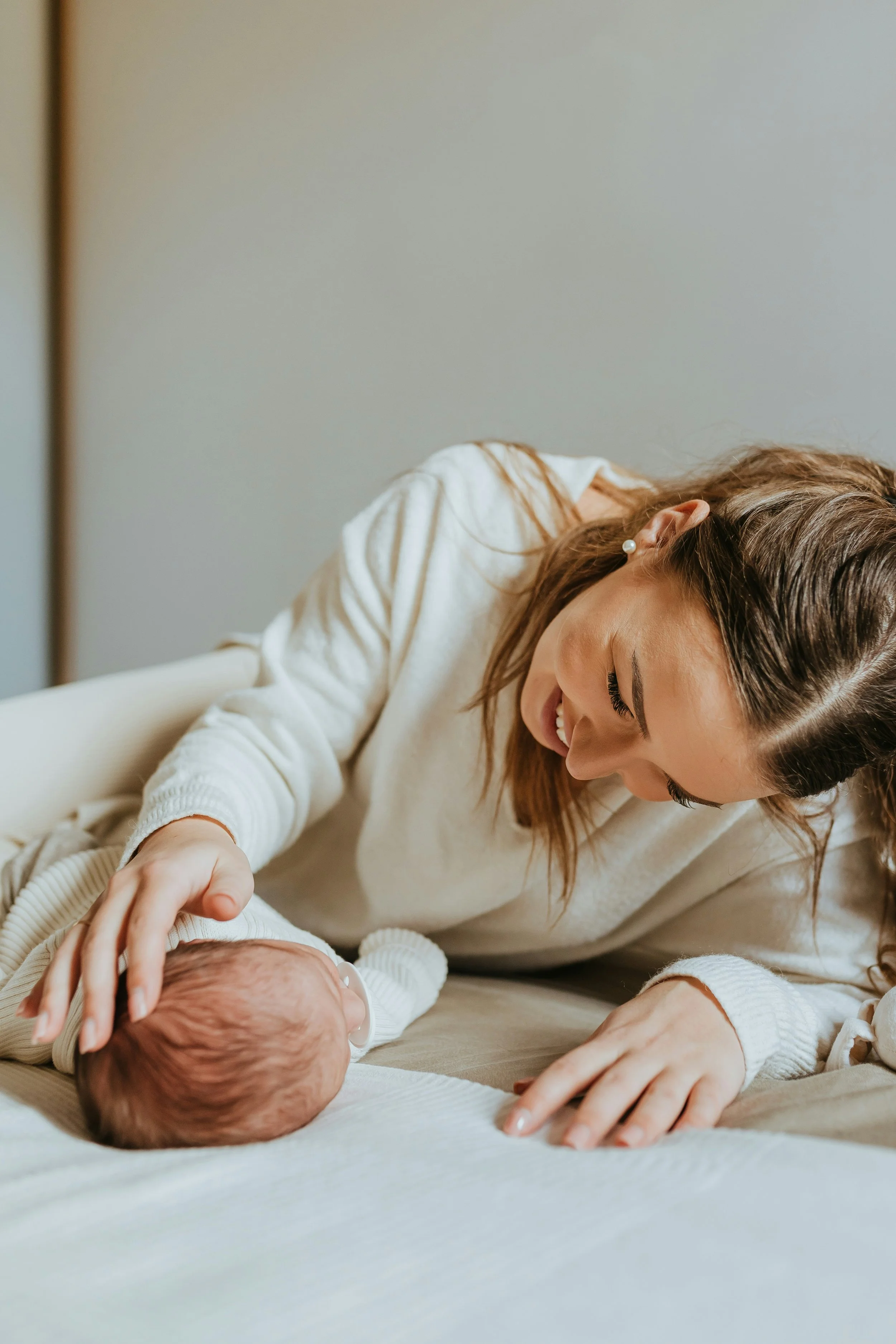 A woman with brown hair, wearing a white sweater, smiling and looking down at a baby, who is lying on their back on a bed, touching the baby's head affectionately.