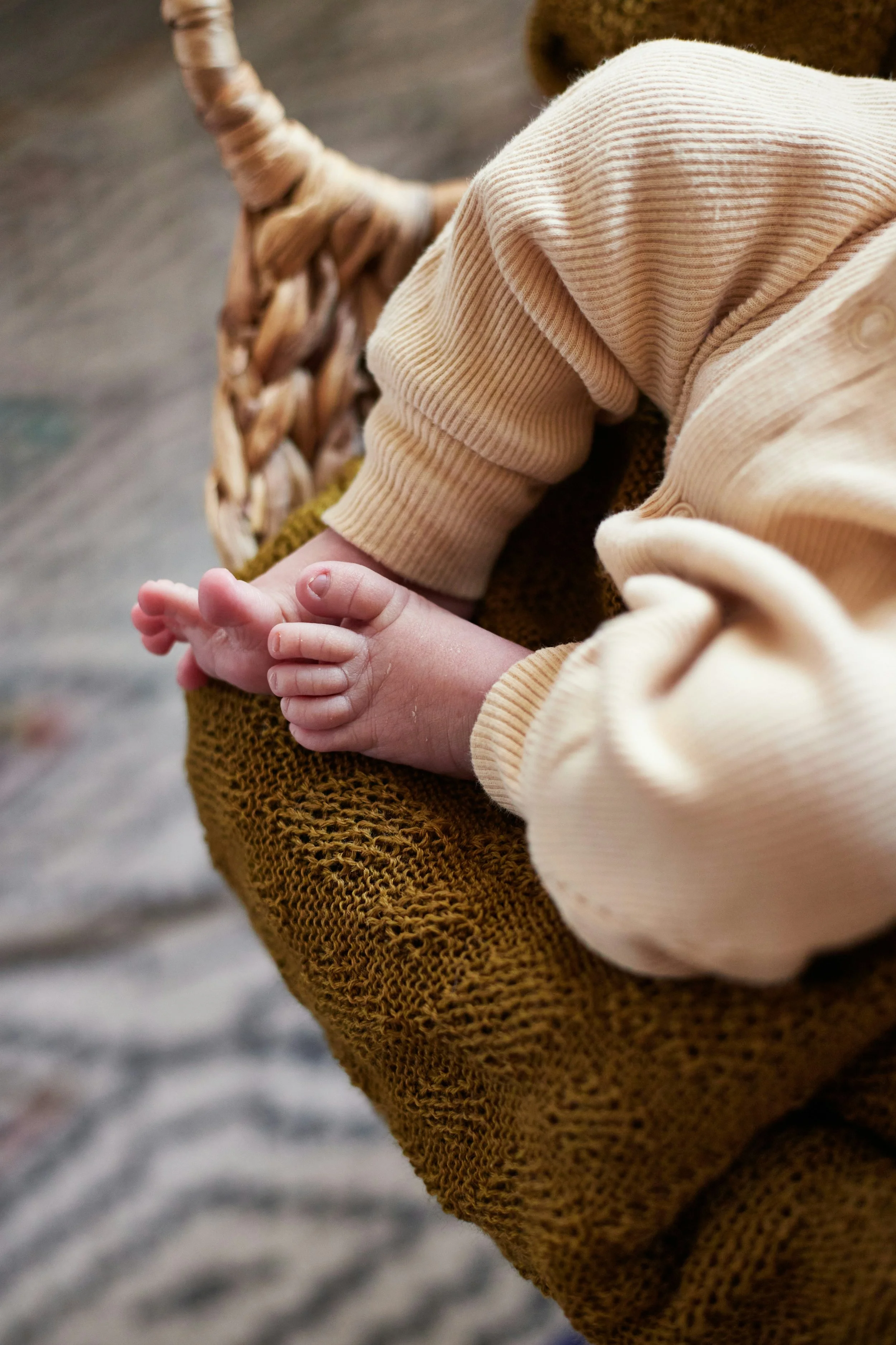 Close-up of a baby's hand resting on a knitted blanket, wearing a cream-colored, long-sleeved ribbed shirt.