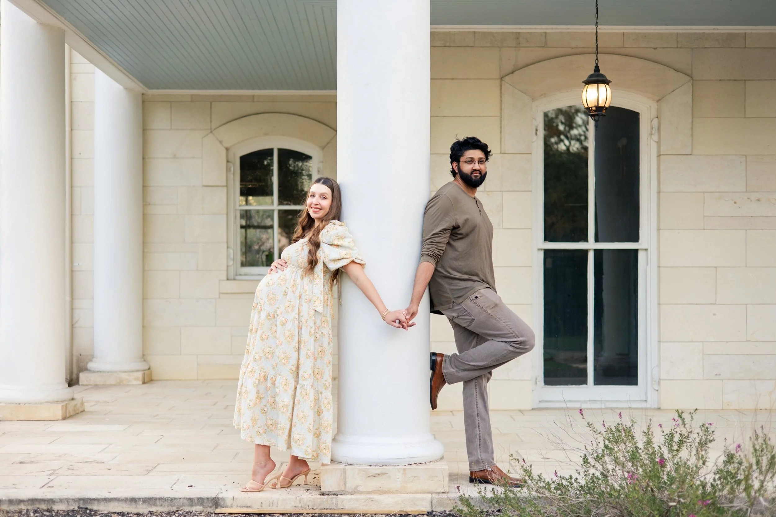 A man and a woman holding hands and leaning against a large white column outside a house. The woman is pregnant, wearing a long floral dress and beige heels, smiling. The man has a beard, wearing a brown shirt and gray pants, with one foot raised and a slight smile. The house has a stone exterior with large windows, a hanging lantern, and some plants in the foreground.