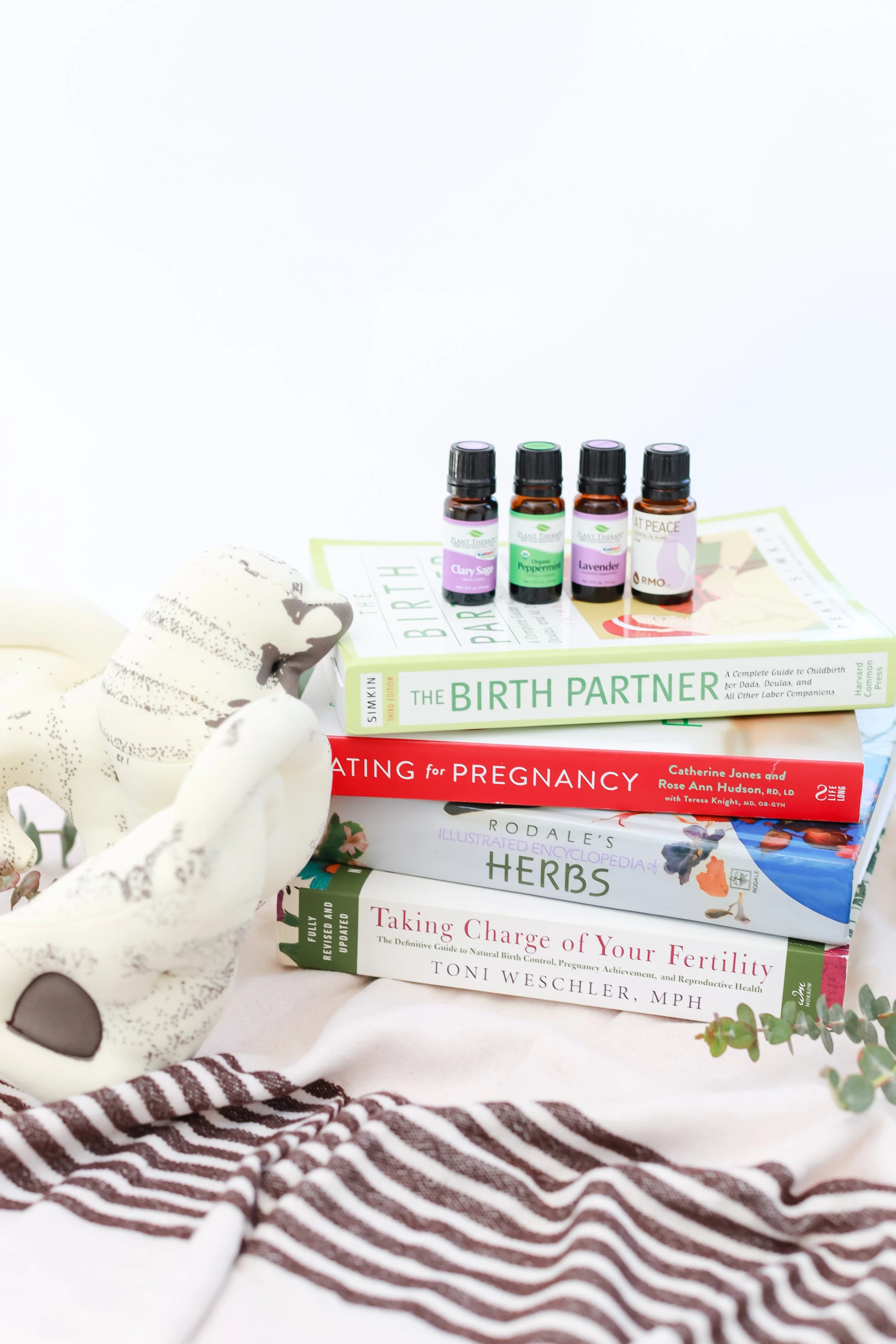 Stacked books about pregnancy, childbirth, and fertility with essential oils and plush toys nearby.
