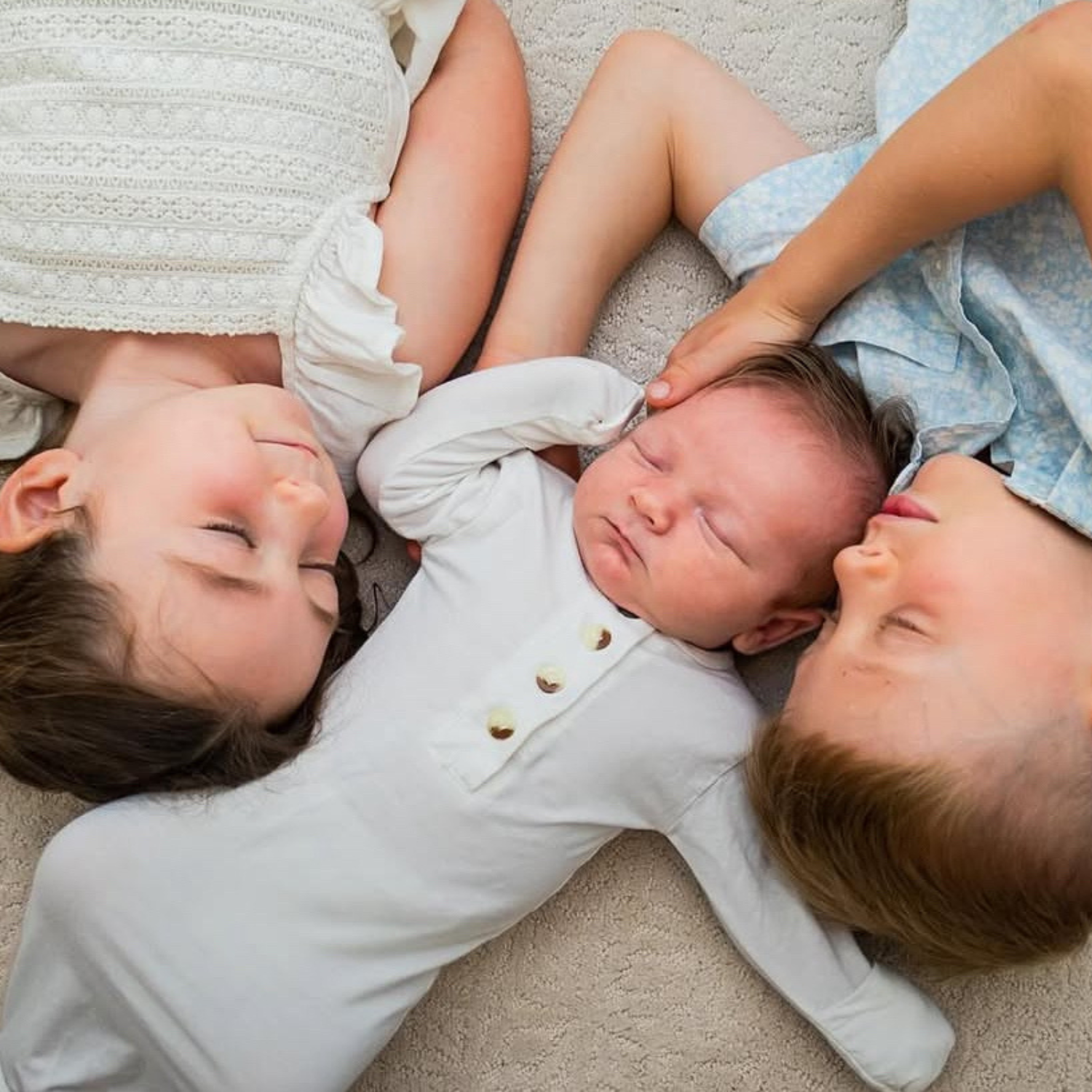 A young girl, a young boy, and a baby lying on a beige carpet with their heads close together, peacefully admiring their new sibling.