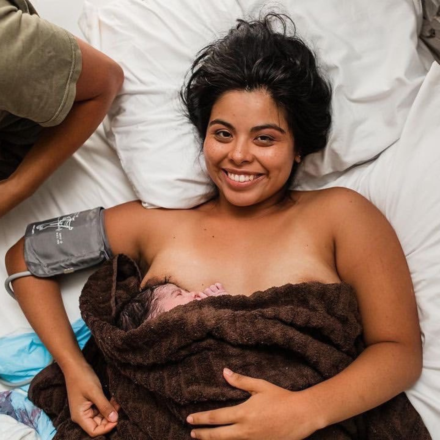 A woman lying in a bed, smiling, with a newborn baby wrapped in a brown towel resting on her chest. She is wearing a blood pressure cuff on her arm and appears happy after giving birth.