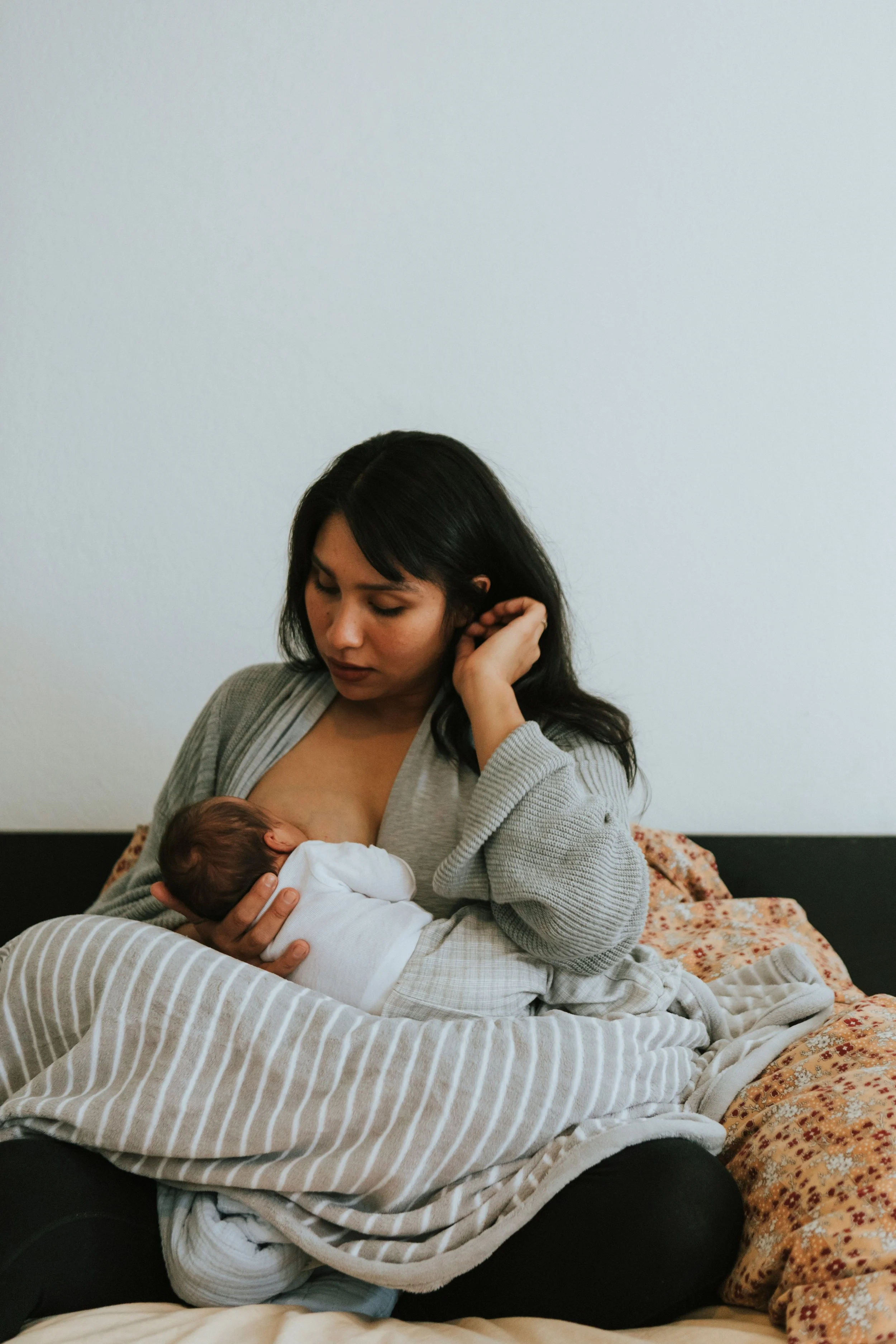 A woman breastfeeding a newborn baby while sitting on a bed with multiple blankets and pillows.