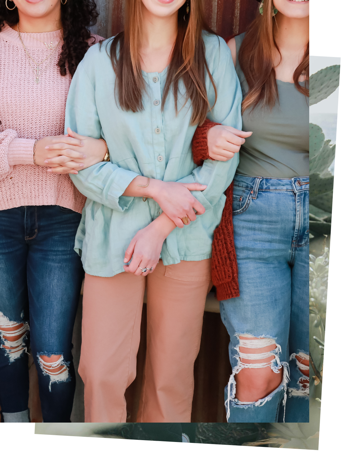 Three women standing side by side, arms linked, wearing casual clothing including distressed jeans, a pink knitted sweater, a blue shirt, and a green top with an orange cardigan, in front of a wooden background.