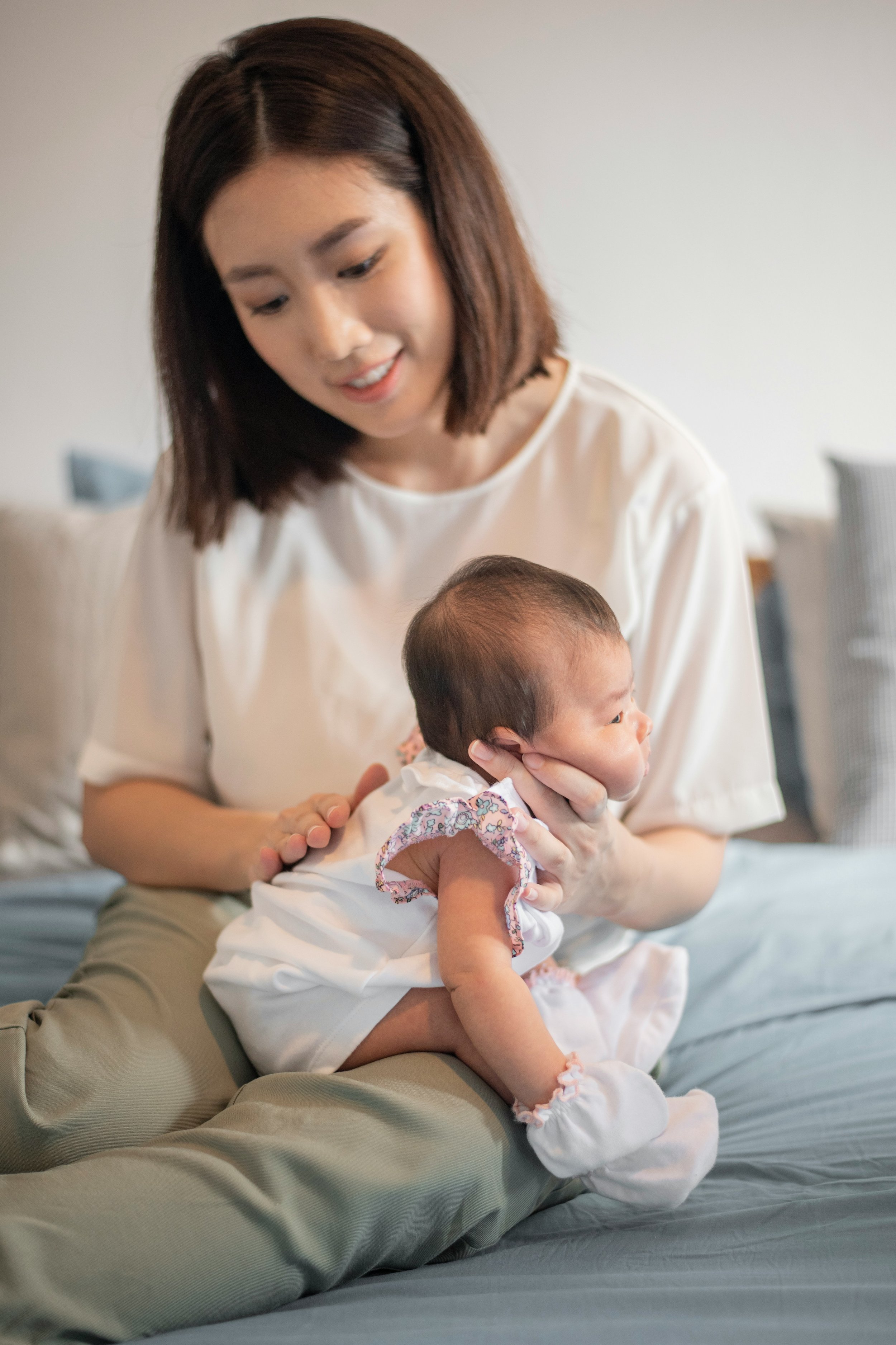 A woman with shoulder-length brown hair playing with a young baby on a bed, holding the baby under the armpits.