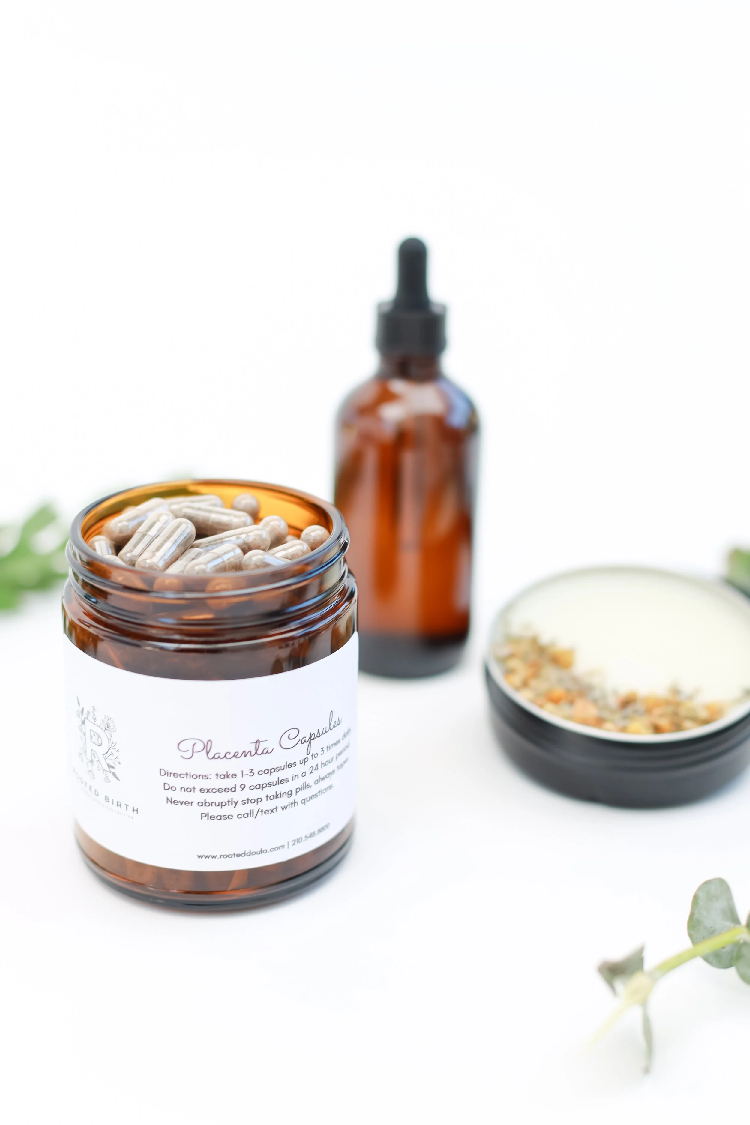 Open amber glass bottle labeled 'Placenta Capsules' with capsules inside, additional amber dropper bottle in the background, and a round container with placenta salve and dried herbs on a white surface.