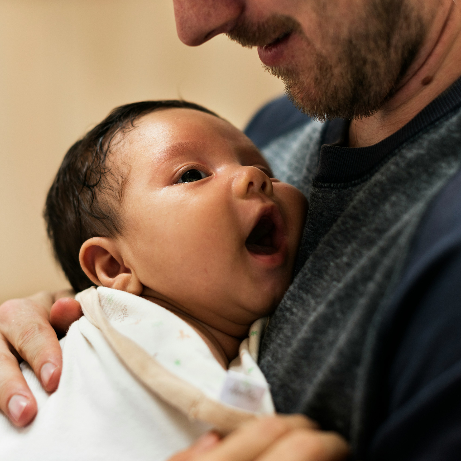 A baby being held close to an adult man's chest, looking up and opening mouth in a moment of interaction or affection.