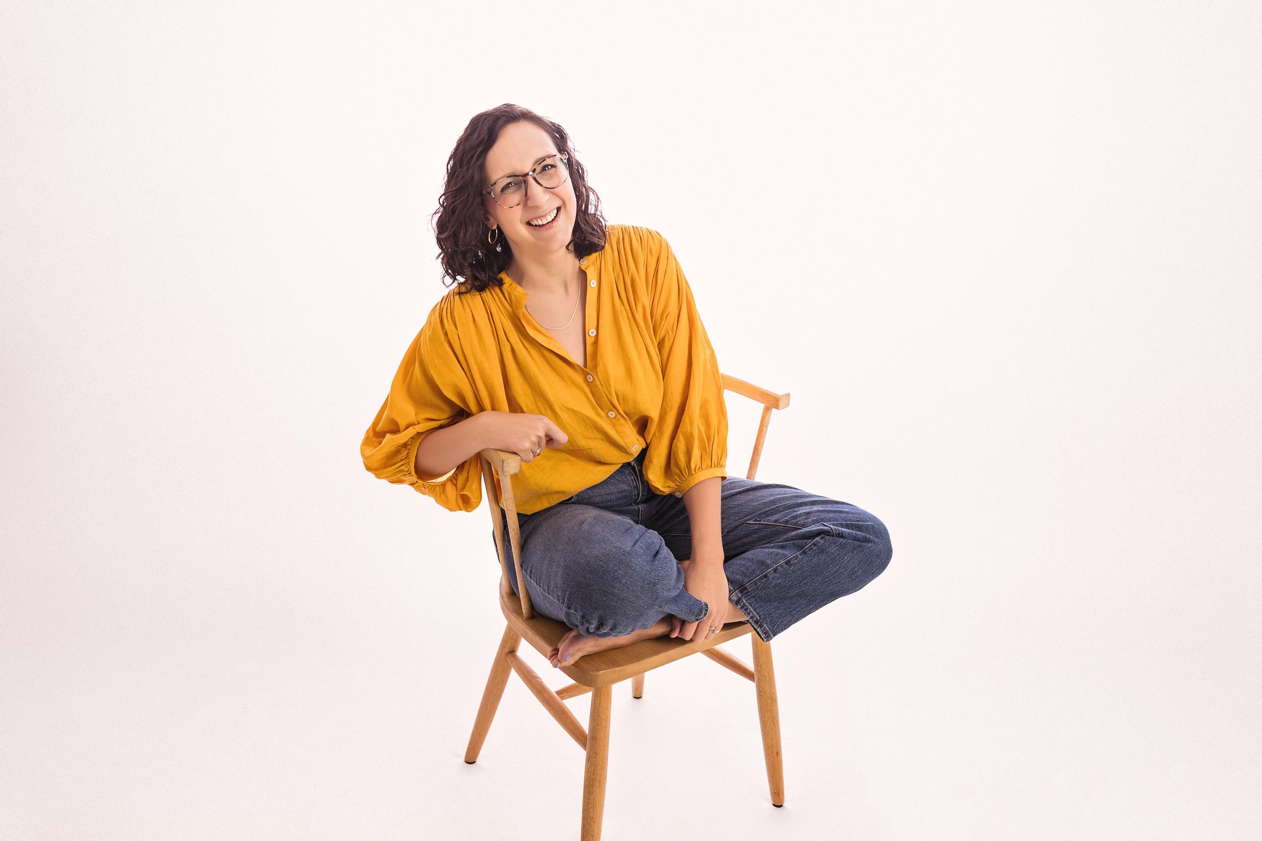 Image of a smiling brunette woman in glasses and a yellow shirt sitting cross-legged in a chair