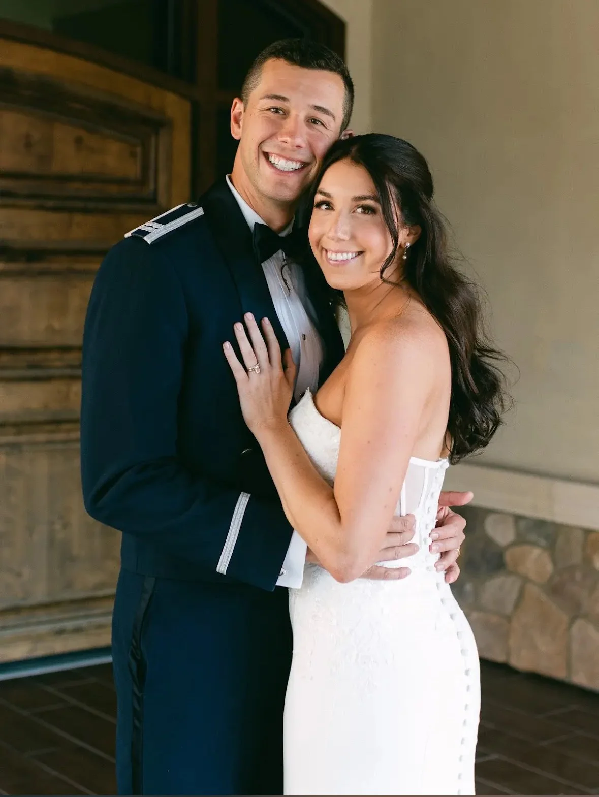 A happy couple on their wedding day, the man in a military uniform and the woman in a white bridal gown, embracing and smiling at the camera.