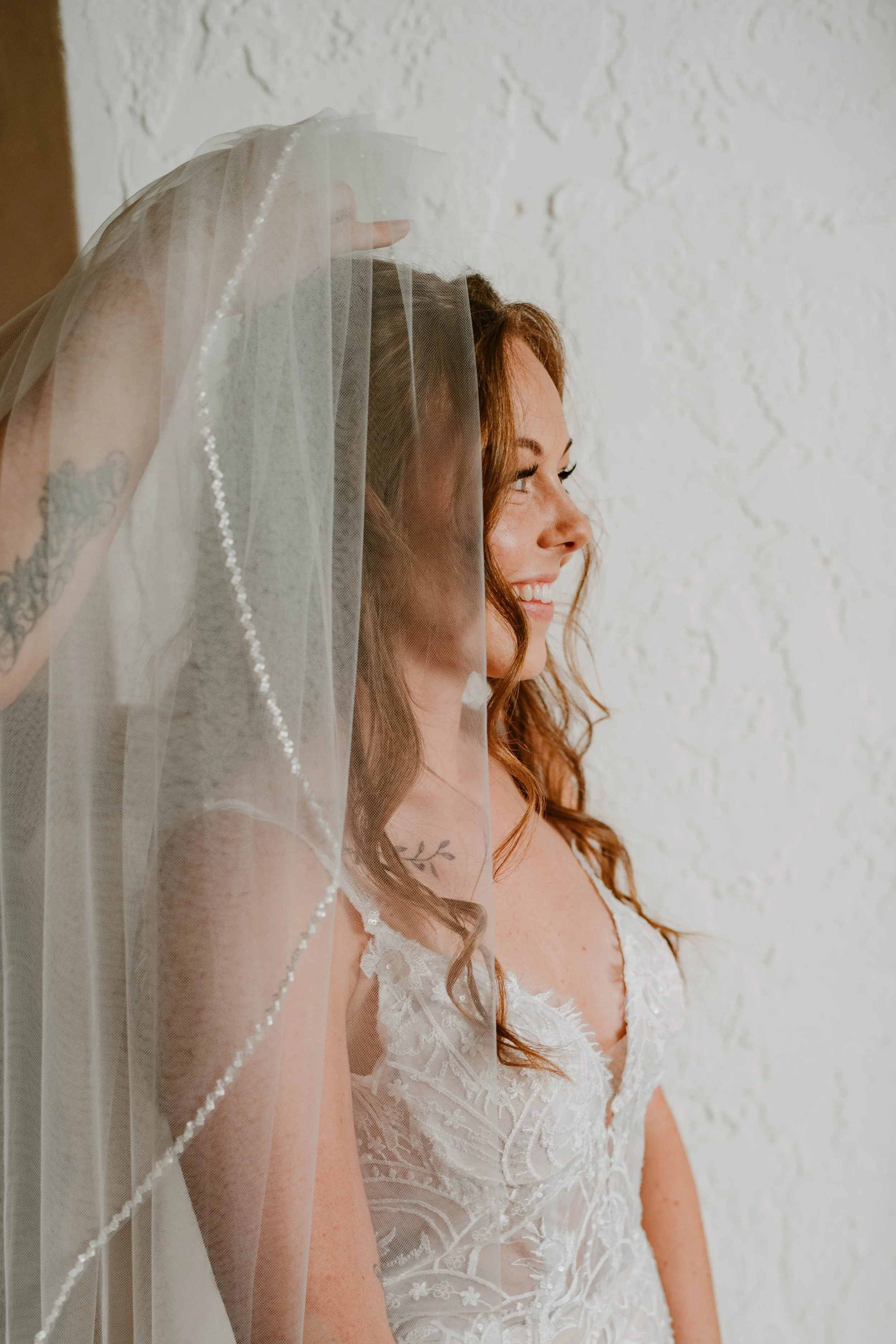 A bride smiling, standing with her veil covering her after getting wedding hair and makeup by marisa andrews