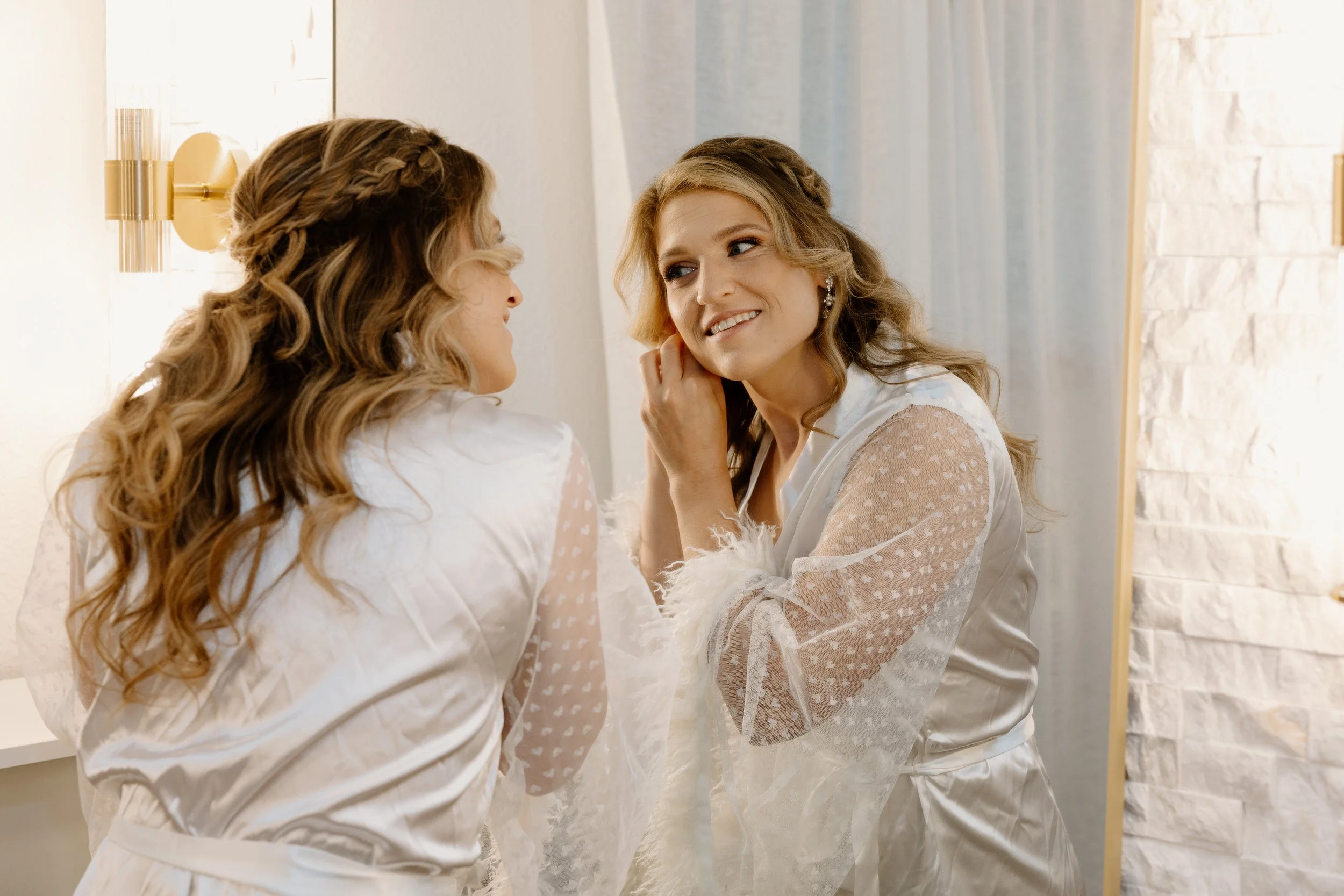 Two women in white satin robes with sheer, polka-dotted sleeves smiling at each other in a well-lit room