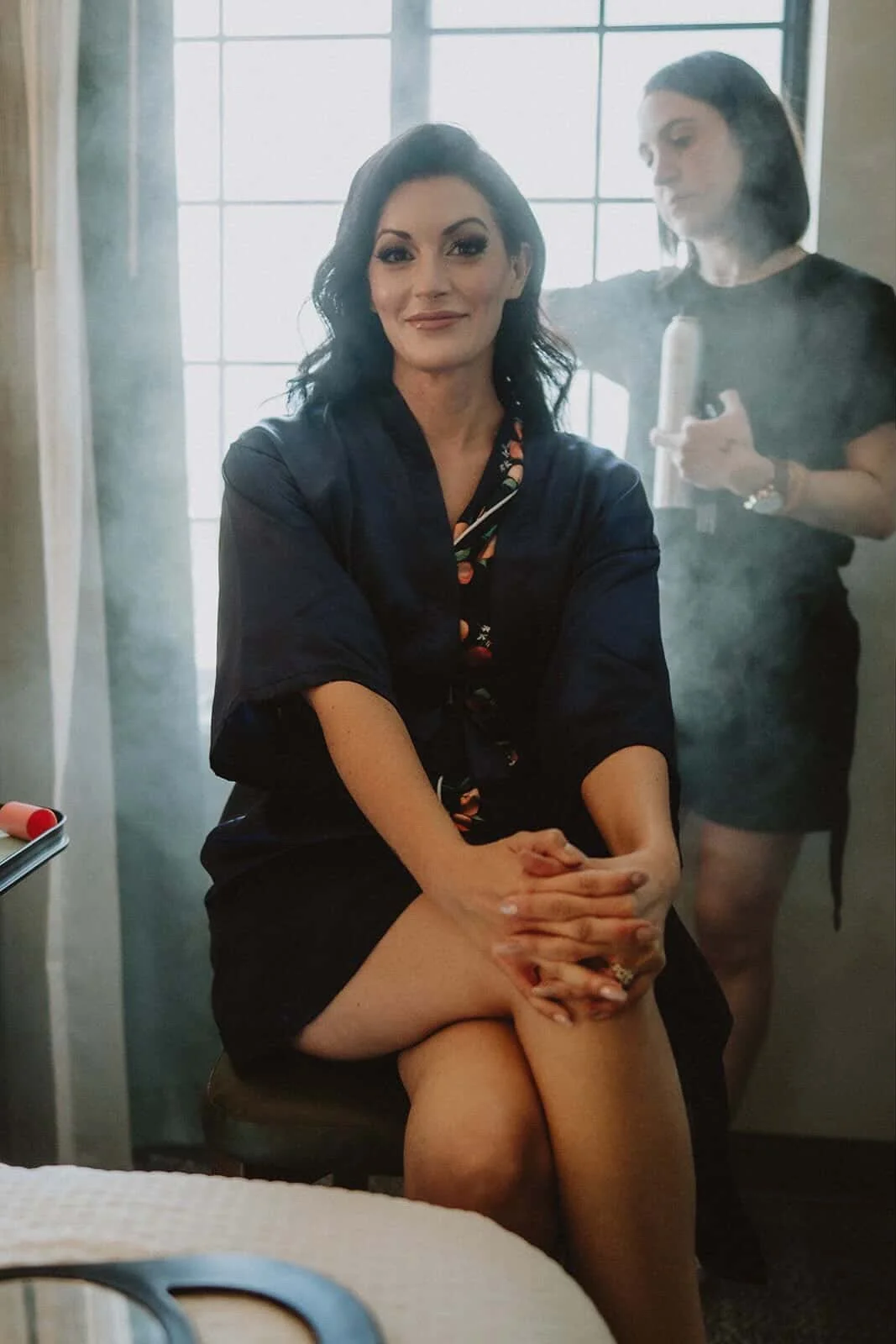 A woman with dark hair sitting on a bed, smiling, with her hands clasped over her lap, while marisa andrews in the background styles her soft glam hair and makeup in a room with large windows.