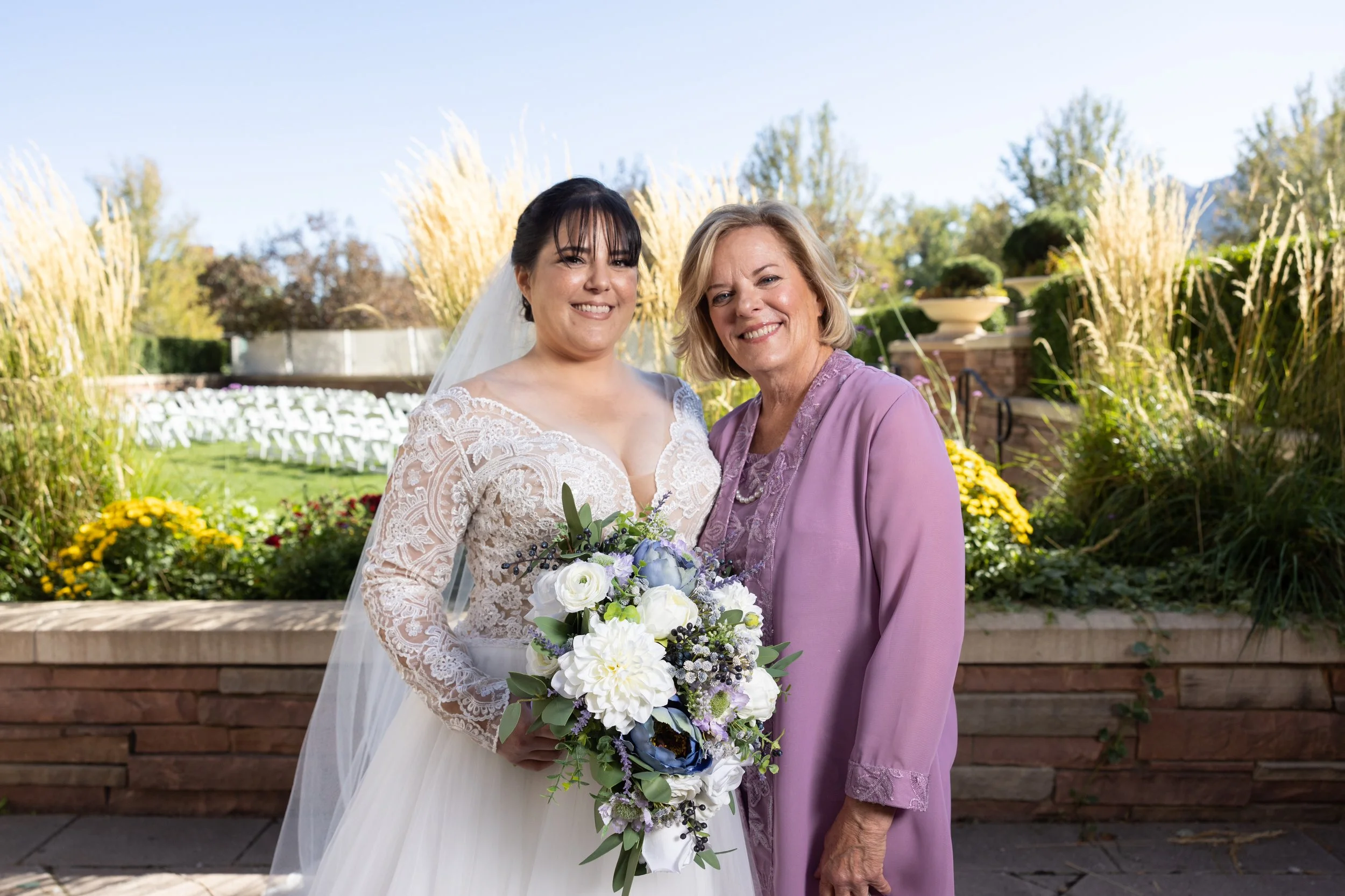 A bride in a white wedding dress holding a floral bouquet standing next to an older woman in a purple dress, outdoors in a garden with greenery and flowers.