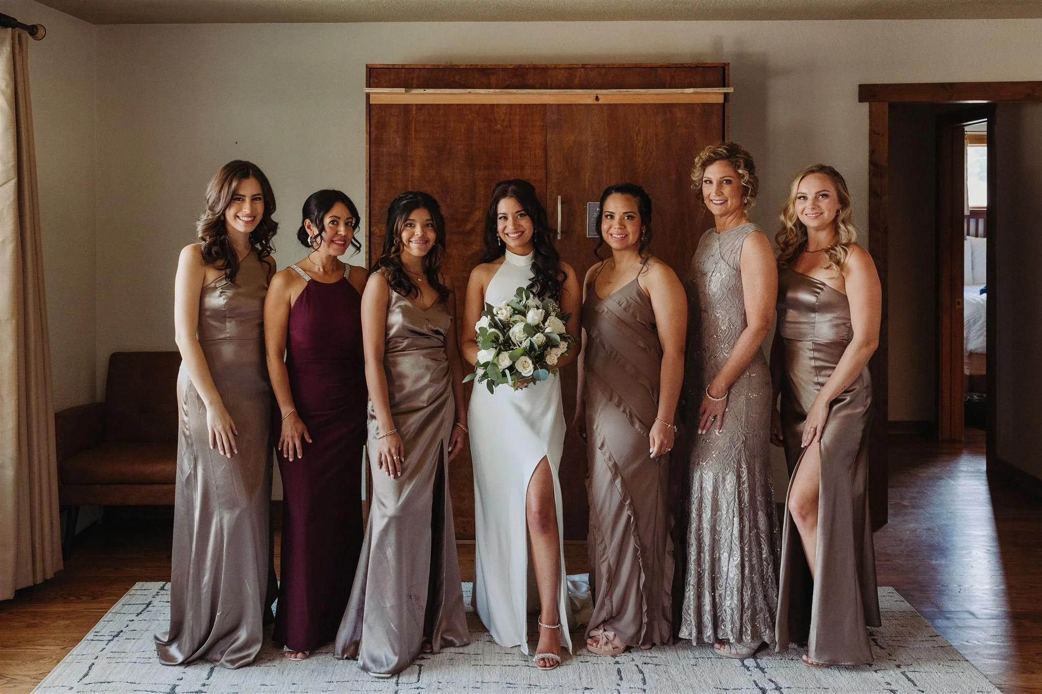 Group of eight women dressed in formal attire, standing indoors, with one woman holding a bouquet of white flowers, likely at a wedding or similar event.