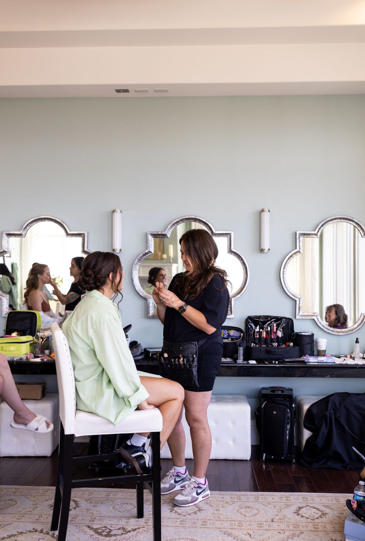 A woman getting her makeup done by a makeup artist in a room with light green walls and decorative mirrors.