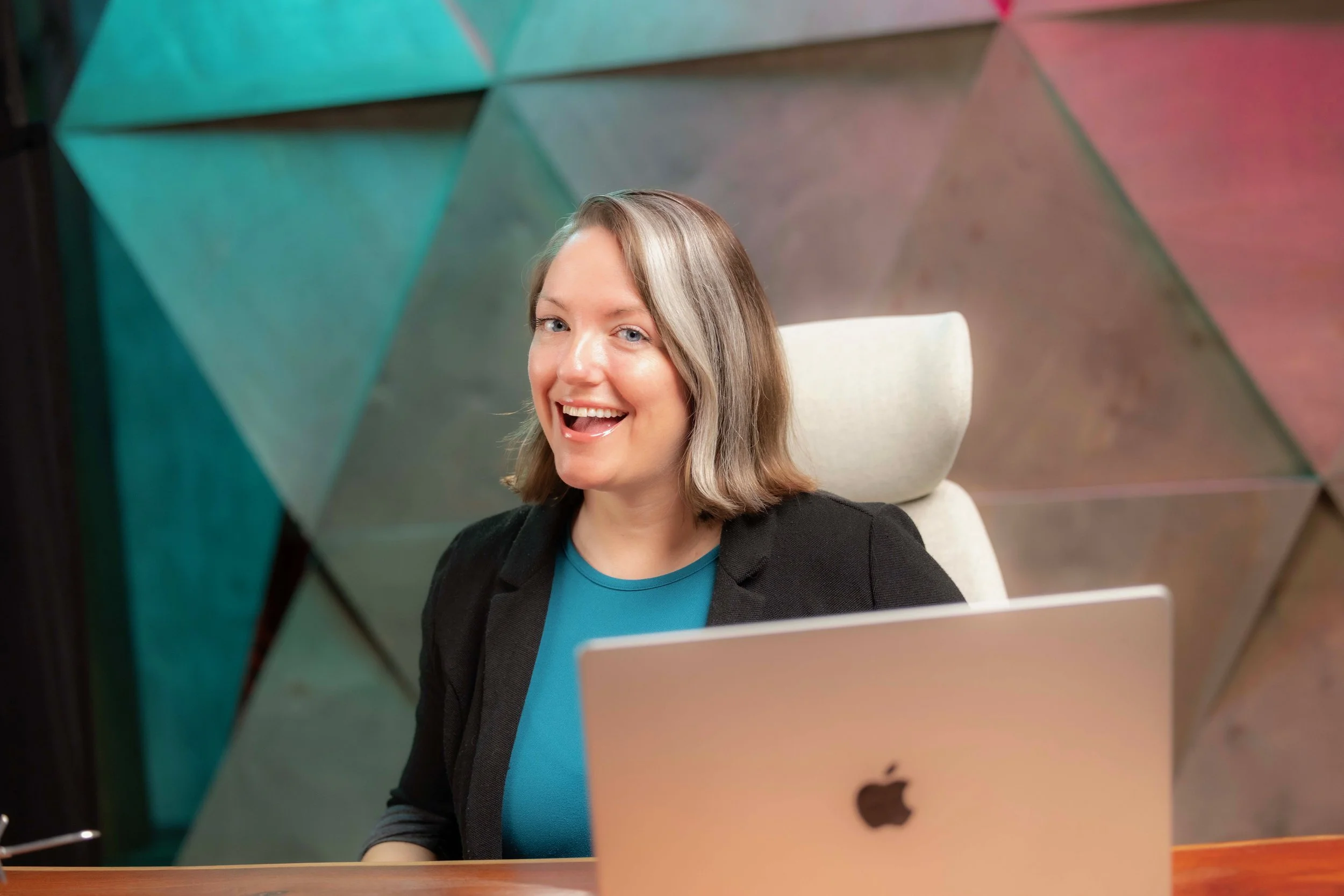 Natalie sitting behind a laptop at a desk with a big open smile