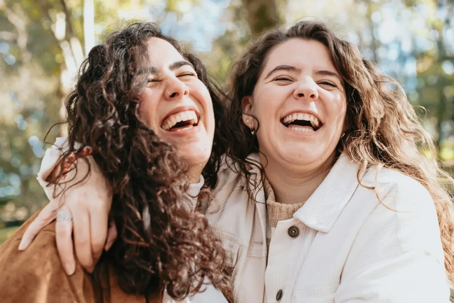 Two women with long brown wavy hair smiling together, one with her arm around the other. The connection and community found in online body image support groups.