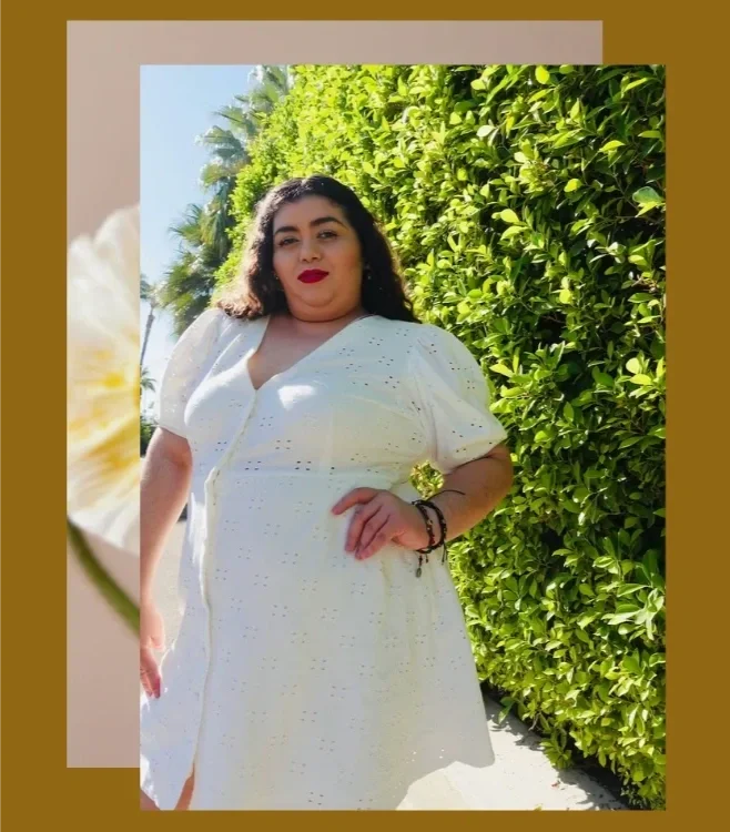 Fat Latinx person with long wavy hair and red lipstick in a white dress against green foliage. Celebrating body liberation and fat-positive joy.