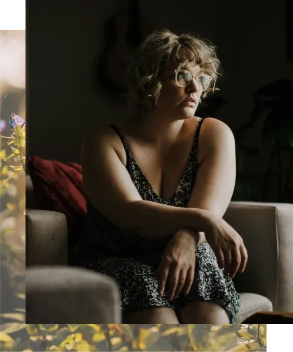Woman with short wavy blonde hair and glasses sitting in a chair, arms crossed, gazing out a window. Therapy for body image and disordered eating in California.