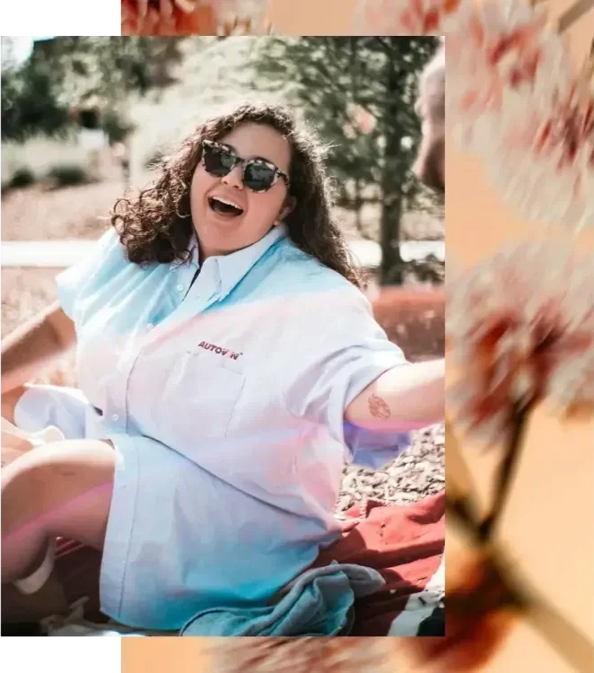 "A joyful fat woman with brown wavy hair and sunglasses smiling broadly against a soft, blurry floral background, embodying body liberation and self-trust.