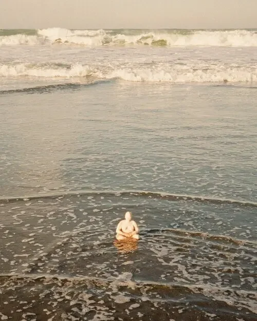 Sculpture of a human body sitting at the ocean with water flowing around it. A symbol of embodiment, resilience, and finding peace in the body.