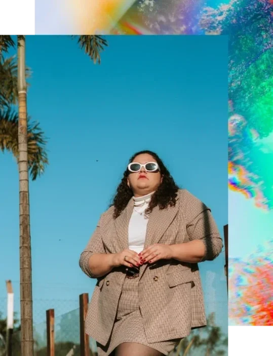 Stylish fat femme with curly hair and white sunglasses posing confidently by a palm tree with rainbow overlay. Body acceptance and HAES therapy in California.