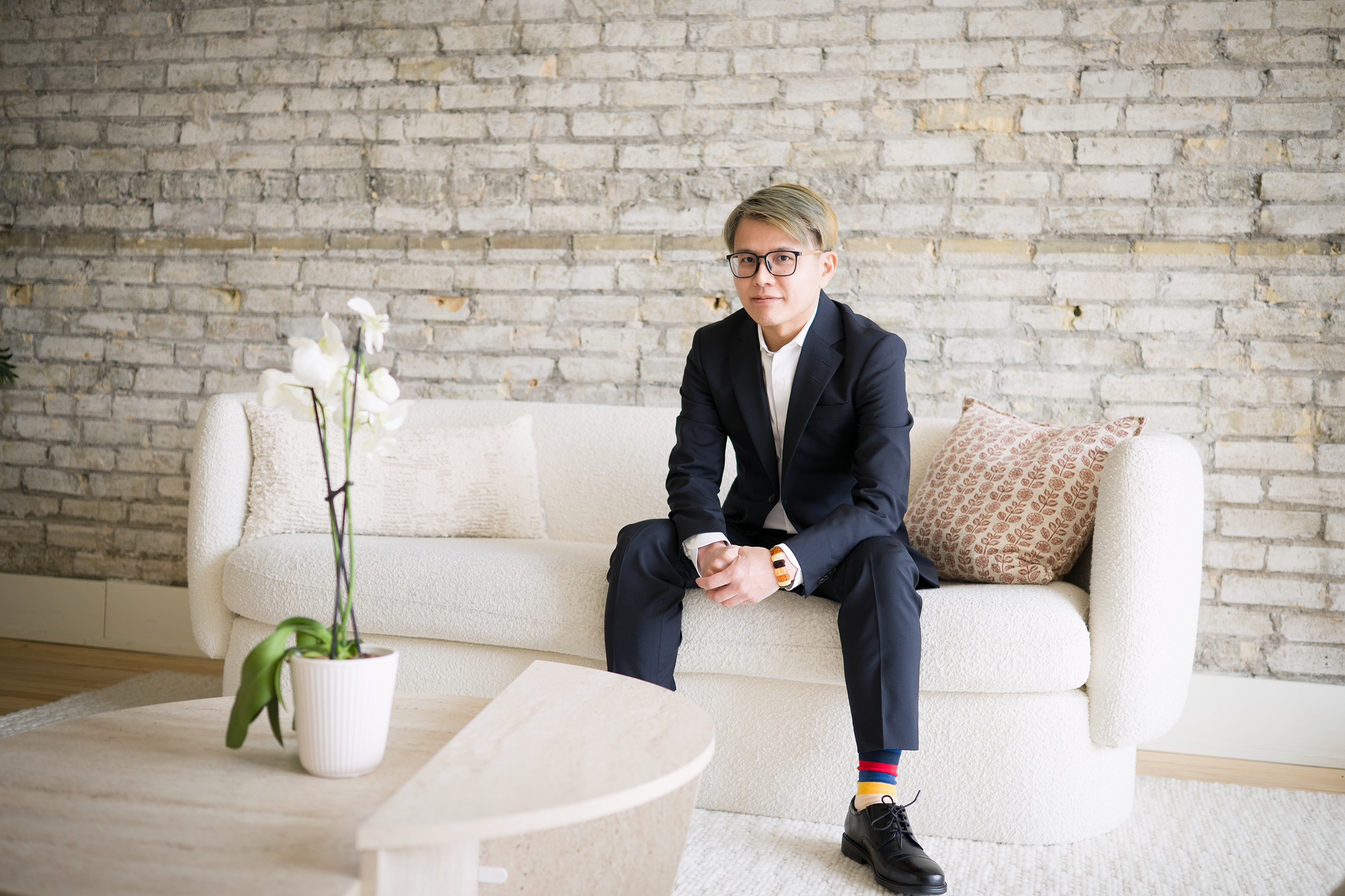 Dr. Chen in a tailored navy suit and glasses sitting on a modern white sofa, posing against a light-colored brick wall in a bright, minimalist interior.