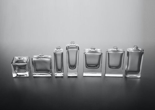 A row of seven glass bottles with varying shapes and sizes, all empty and clear, placed on a reflective surface against a plain background.