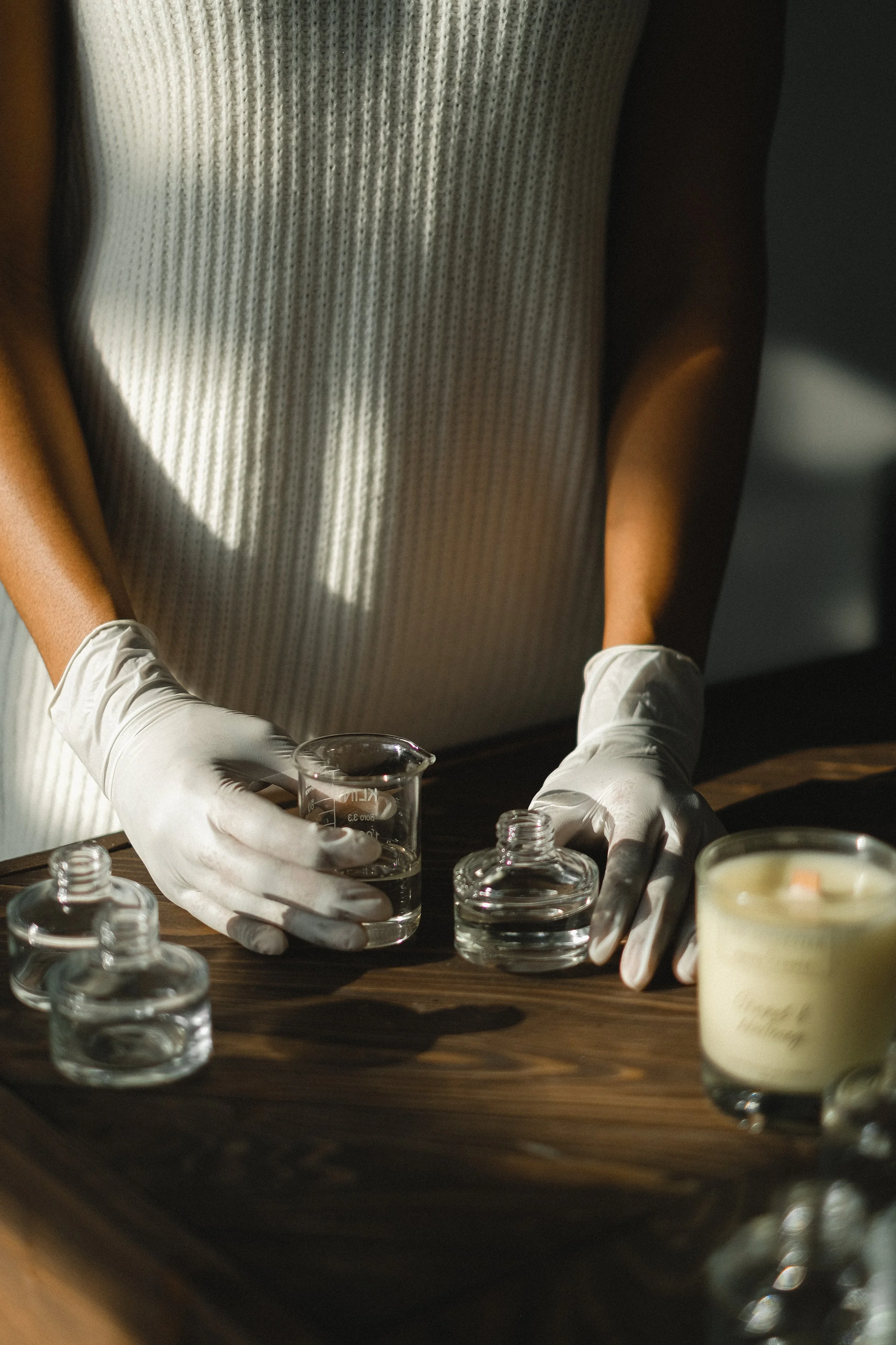A person in a light-colored top wearing white gloves is handling glass laboratory equipment, including beakers and small bottles, on a wooden surface. There is a candle on the right side of the image.
